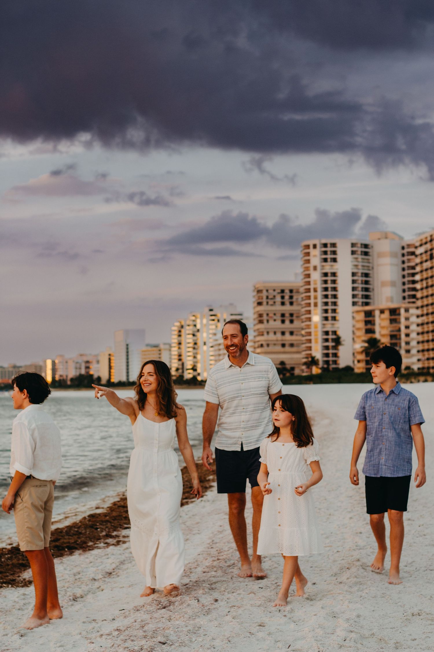 Group of people walking together along a white sandy beach at sunset with coastal buildings in the background.