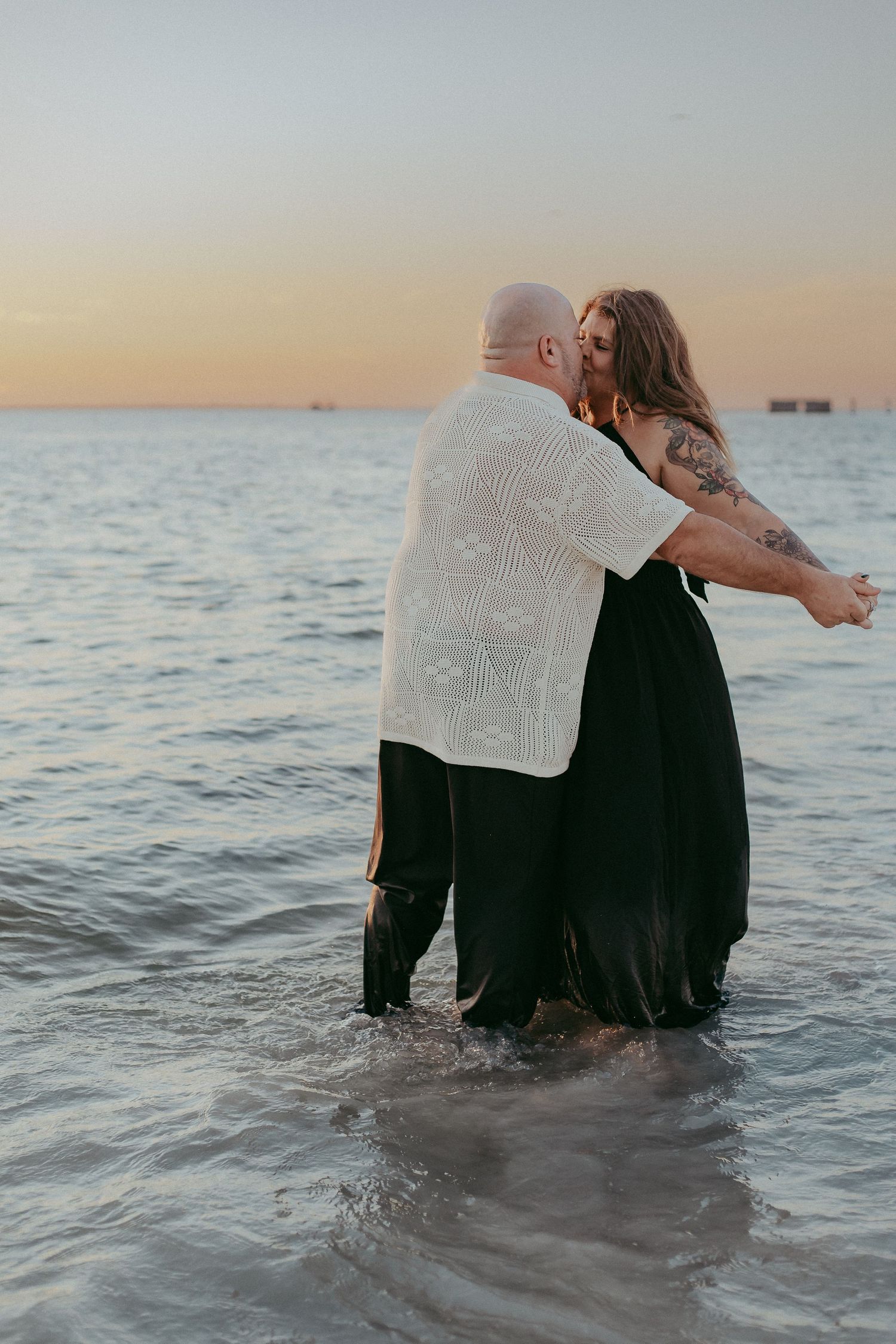 A couple sits together in ocean waves at sunset while reading a newspaper, creating a romantic beach scene.