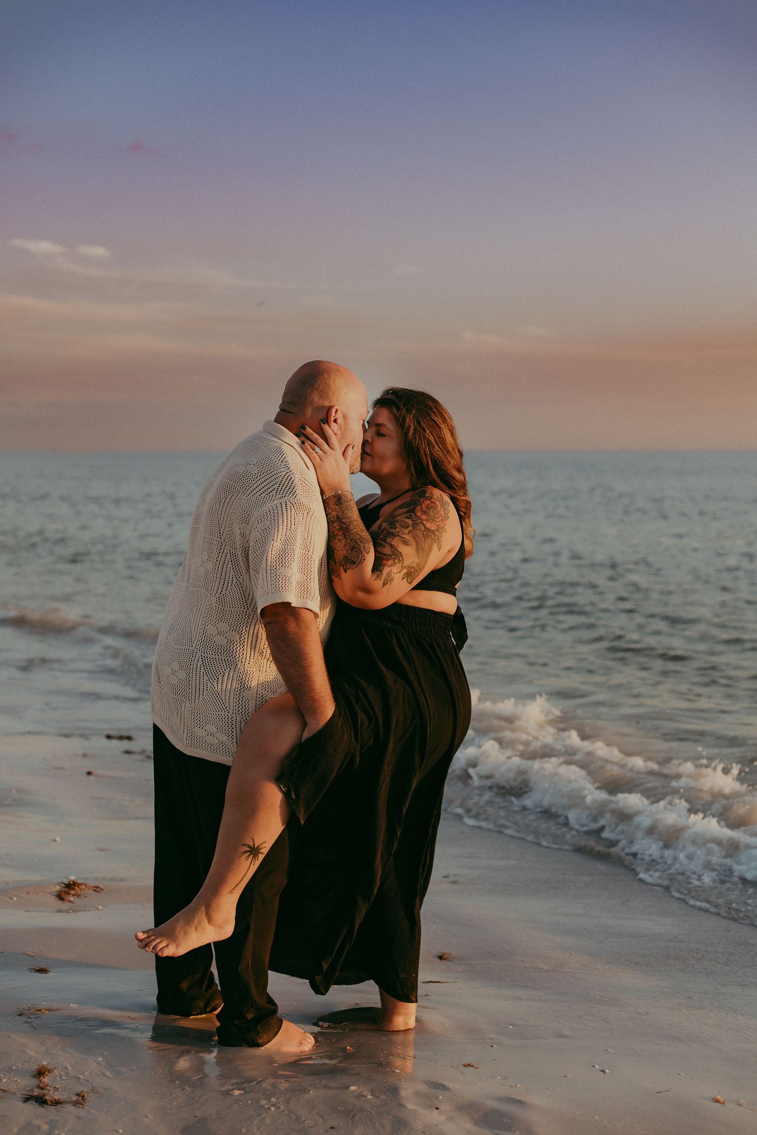 A couple shares a romantic moment on the beach at sunset as gentle waves roll in along the shoreline.