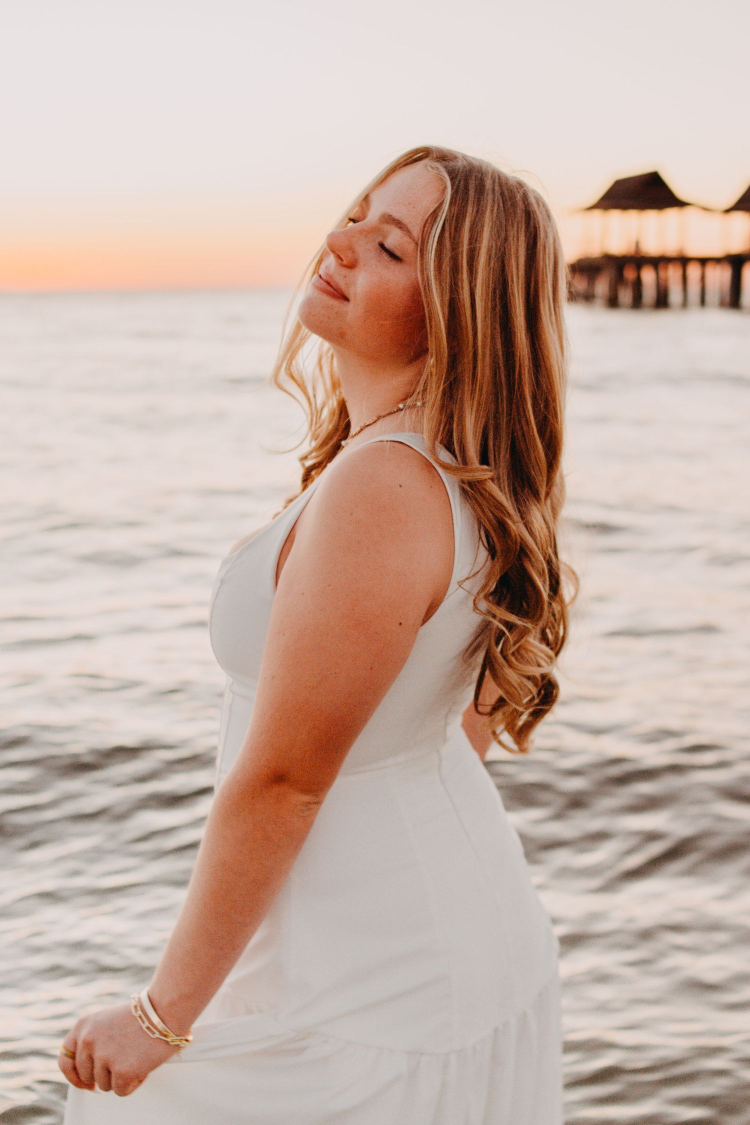 A person in a white dress stands by the ocean at sunset with overwater bungalows visible in the background.