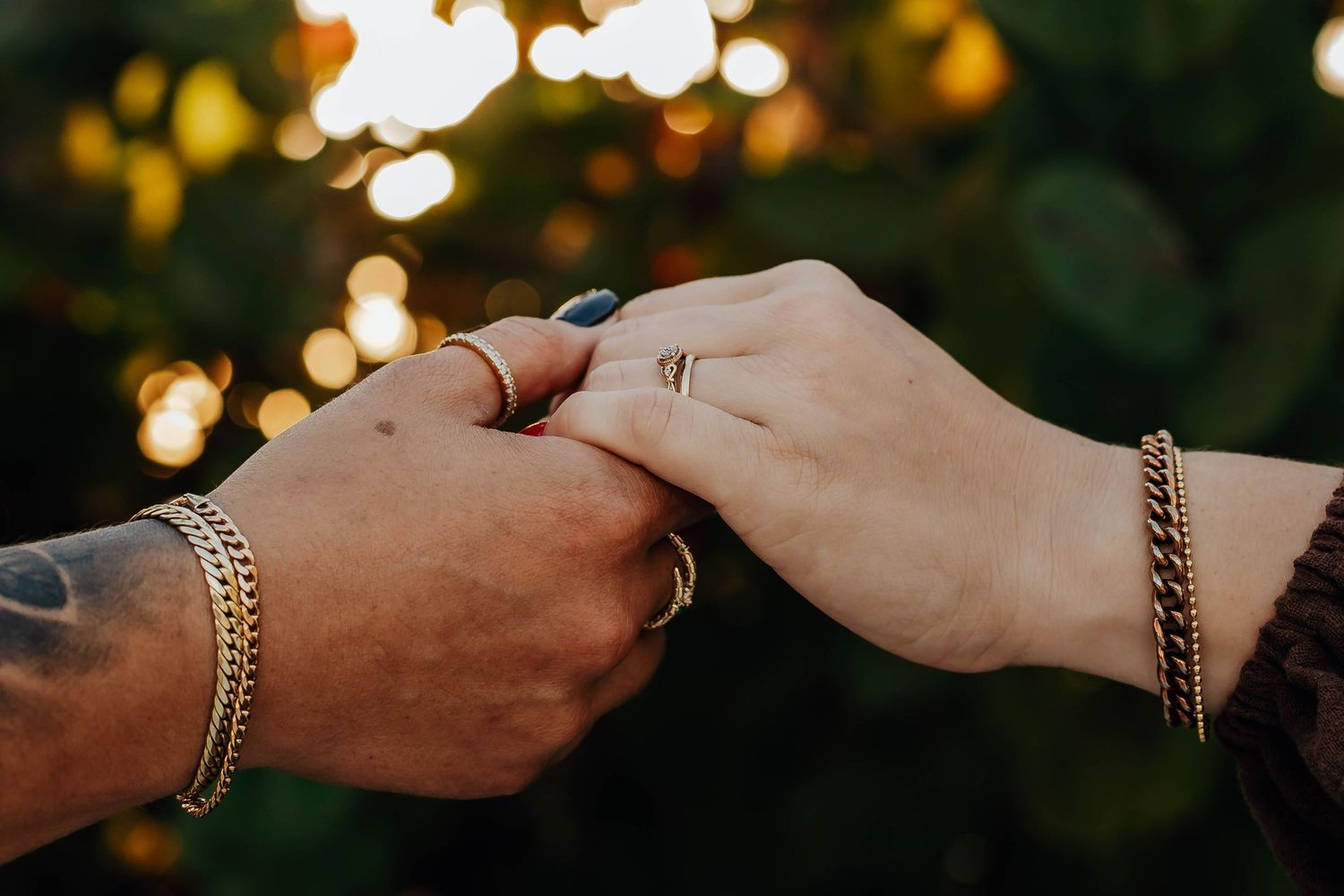 Two people holding hands with gold bracelets and rings, with bokeh lights in the background.