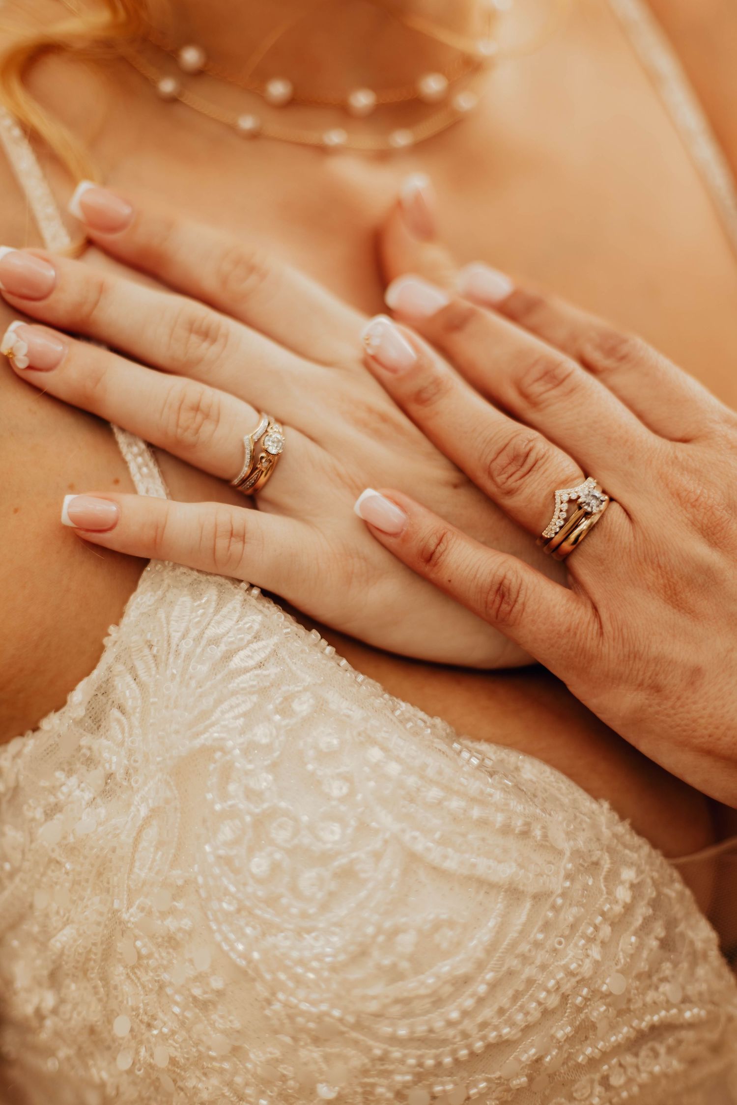 Close-up view of hands with engagement ring resting on delicate white beaded lace wedding dress fabric.