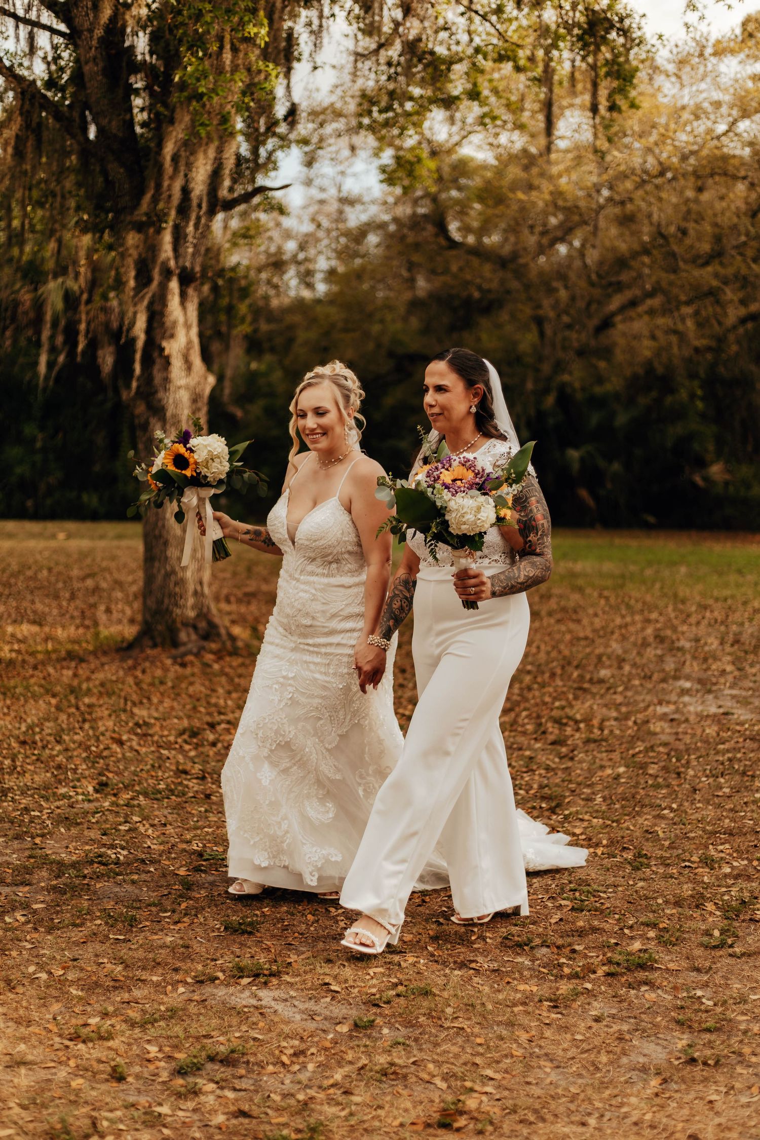 Two brides in white wedding dresses walk hand in hand through autumn leaves in a rustic outdoor setting.