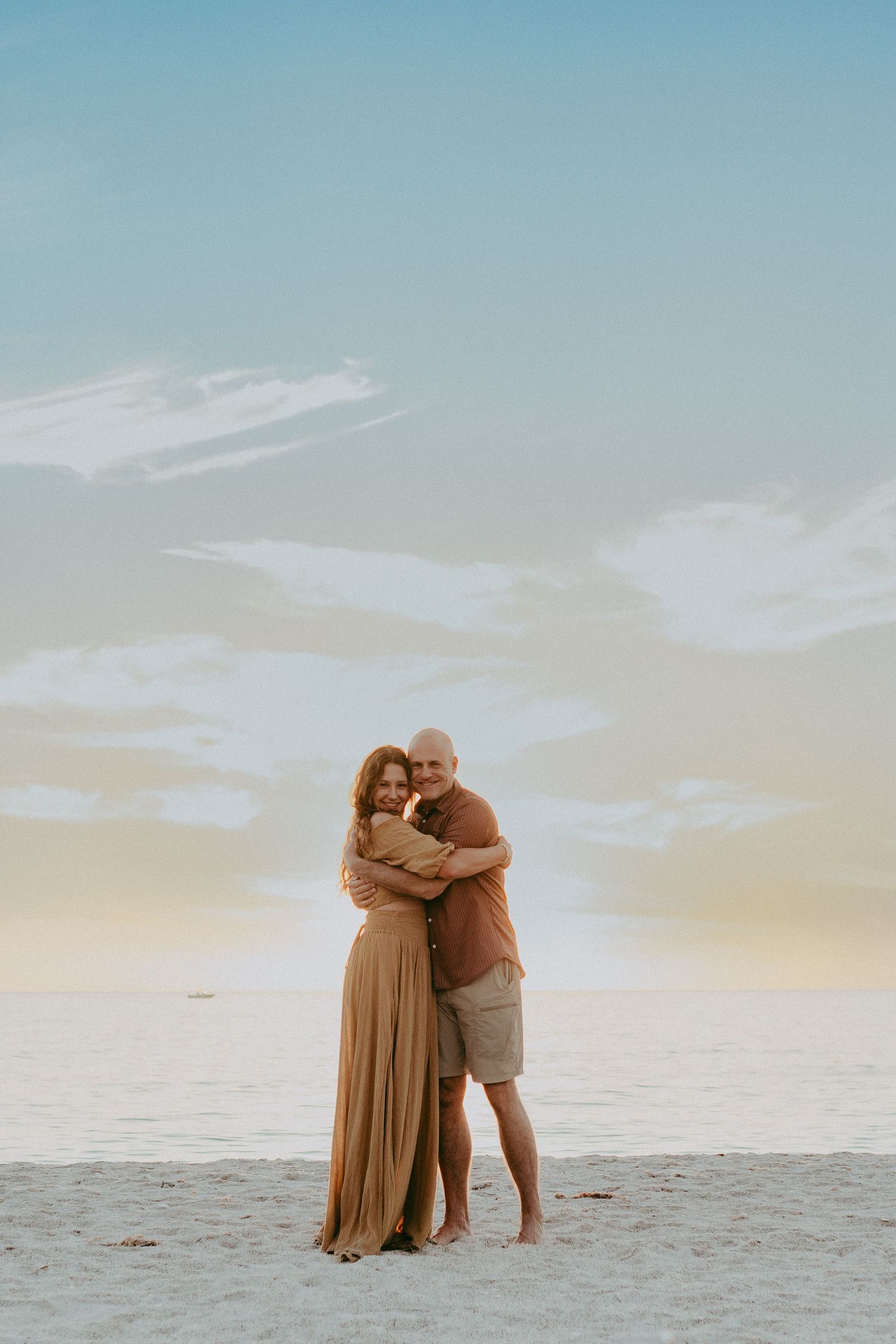 Two people embrace while standing on a sandy beach at sunset with soft pastel sky in the background.