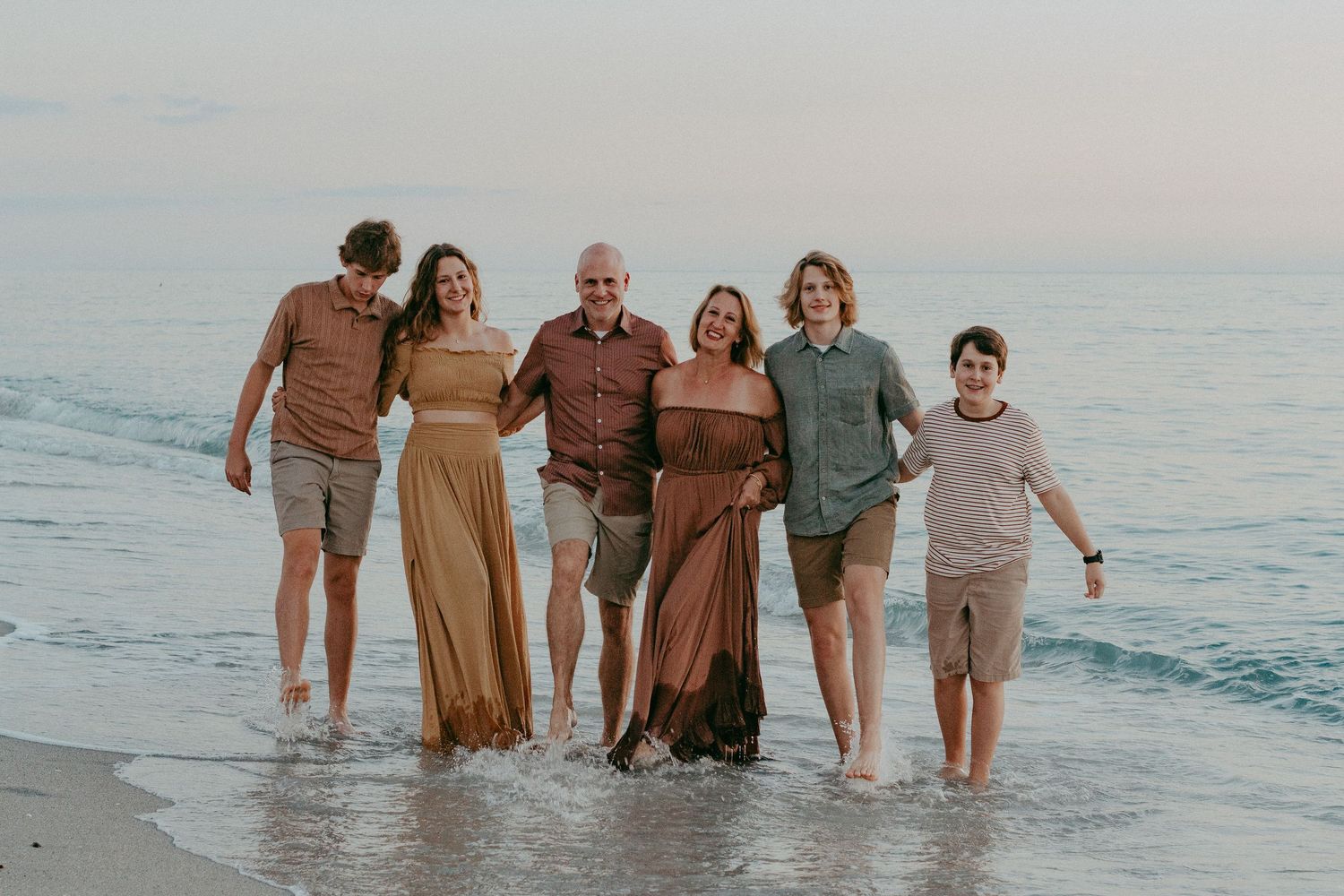 A group in coordinated earth-tone outfits stands together smiling at the beach during sunset.