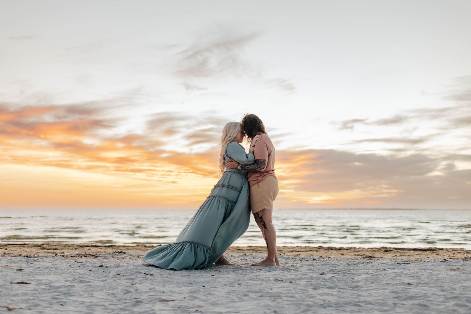 Couple embracing on sandy beach at sunset, woman in flowing teal gown, romantic oceanside portrait.