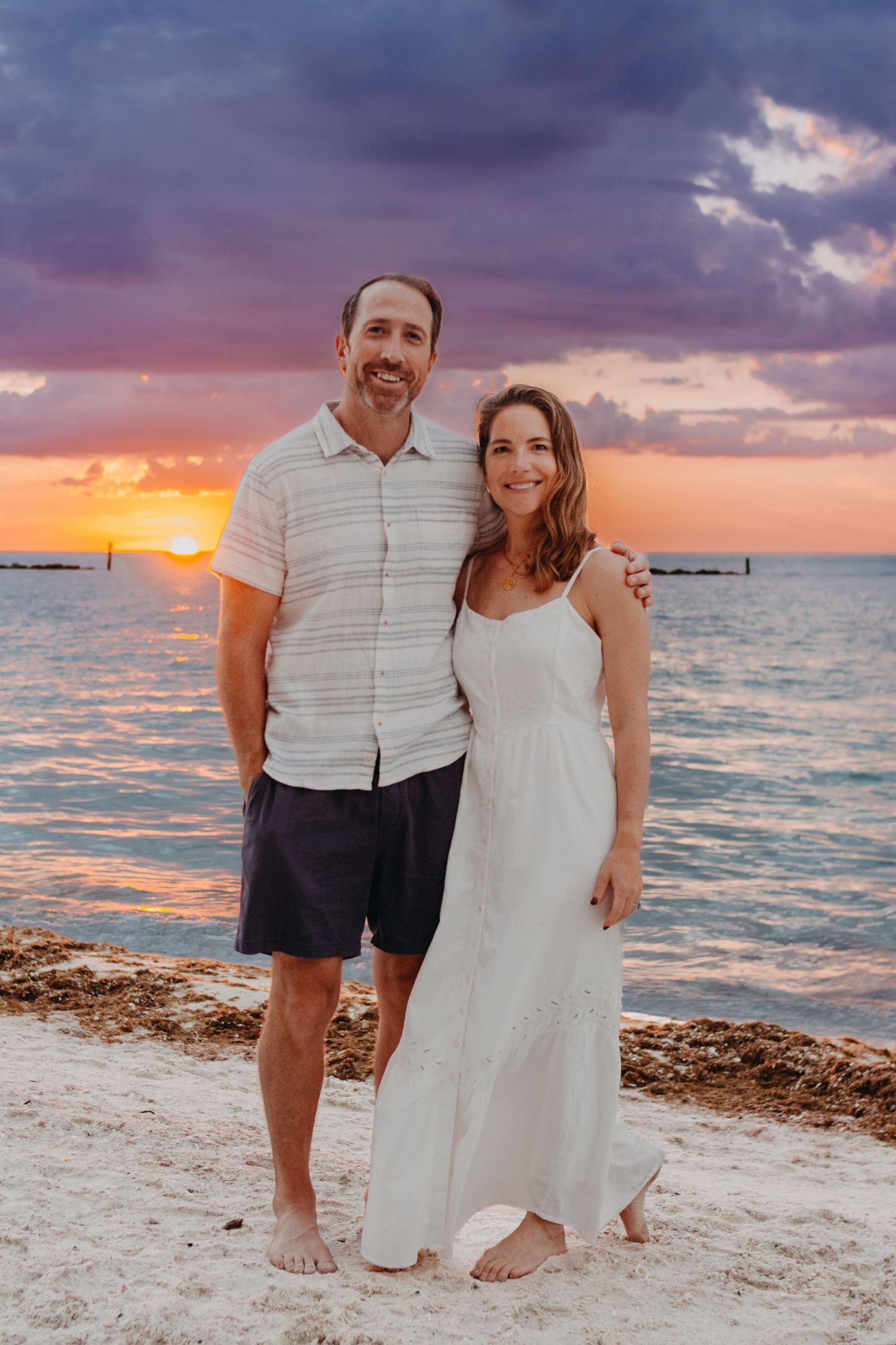 A couple poses together on a sandy beach at sunset with dramatic purple and orange clouds over the ocean.