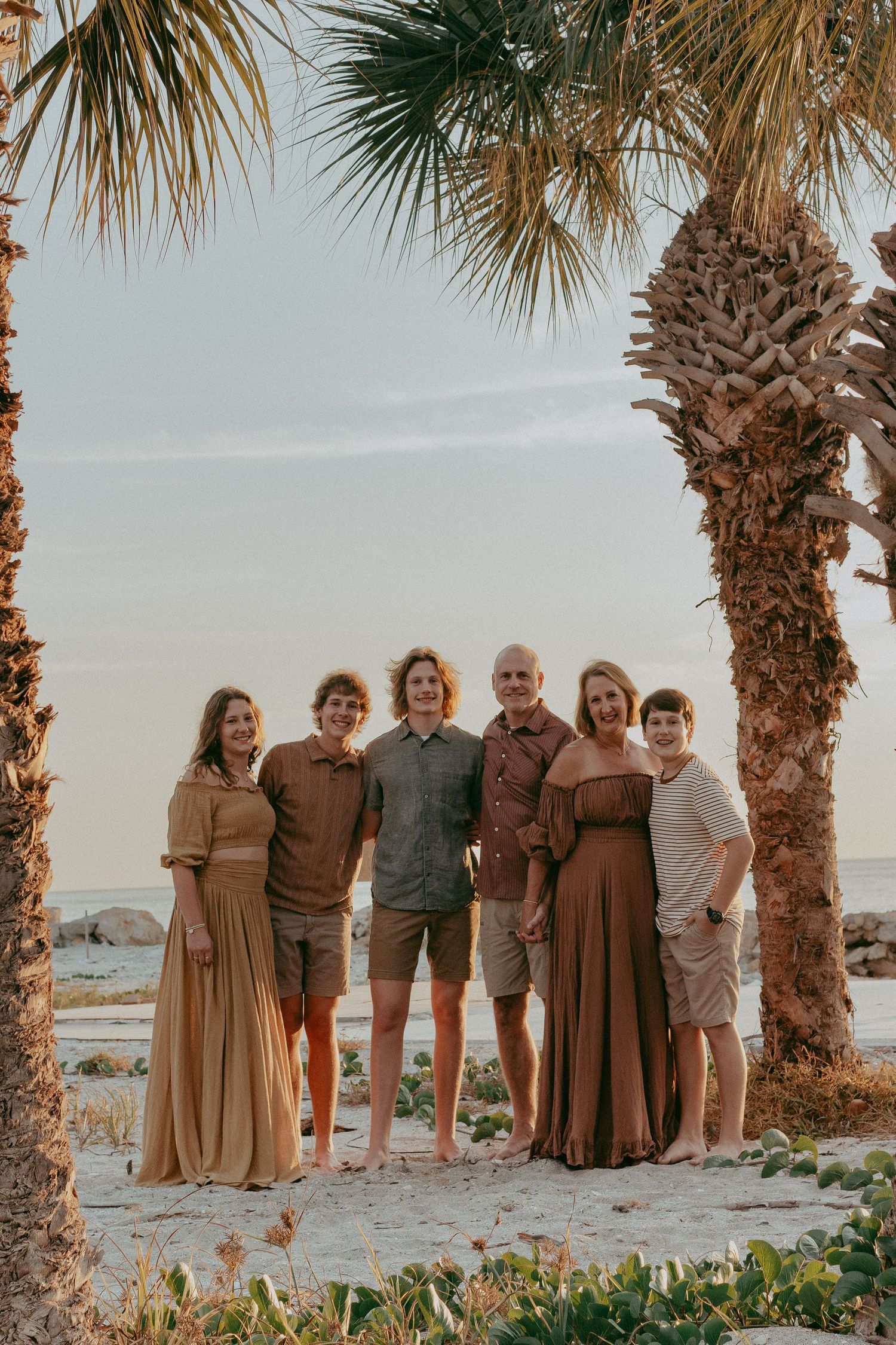 A group stands together on a beach with palm trees and sand dunes at sunset in warm brown and neutral tones.