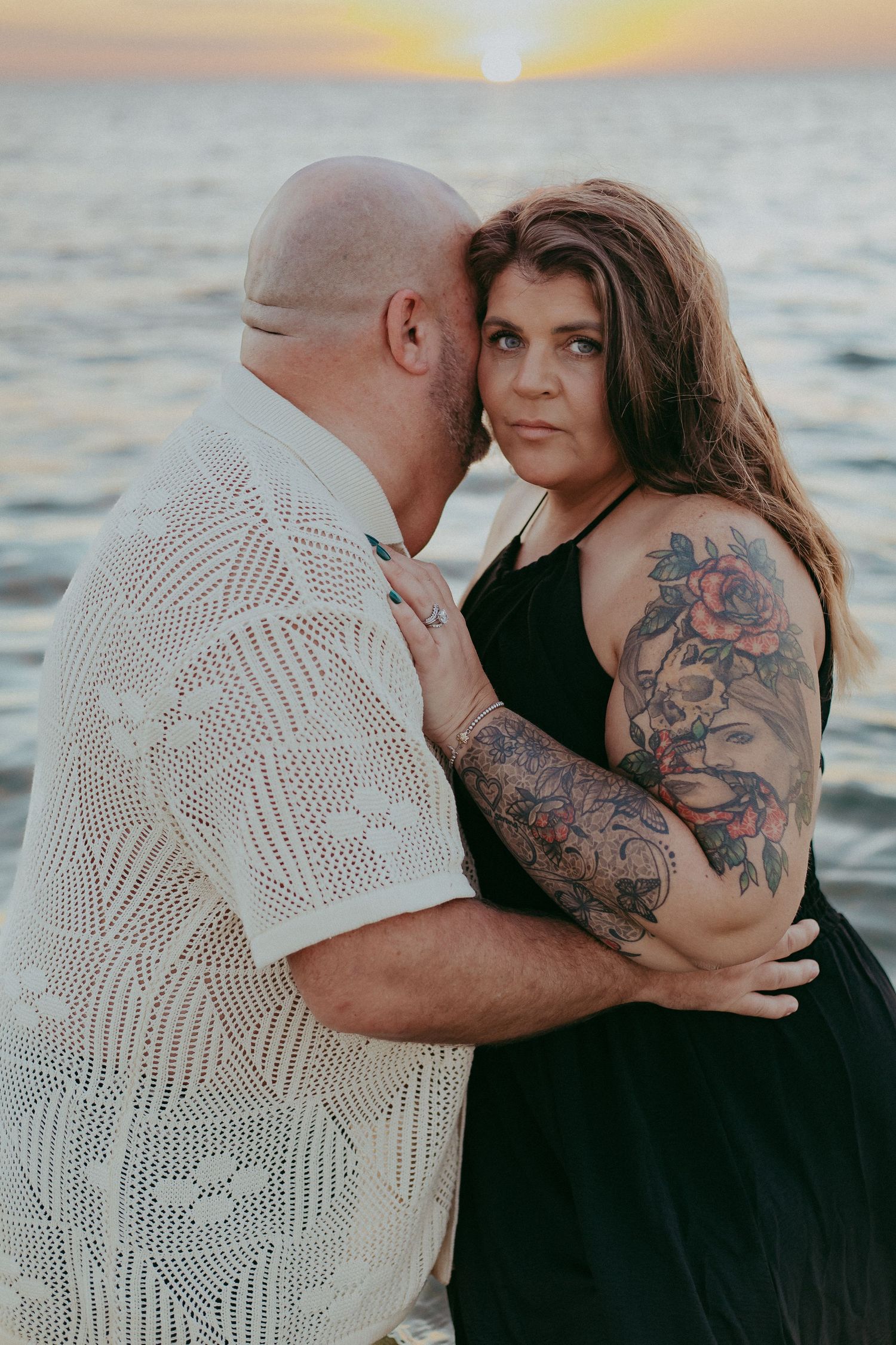 Romantic couple embraces at sunset by the ocean, with visible tattoos and a black dress against white shirt.