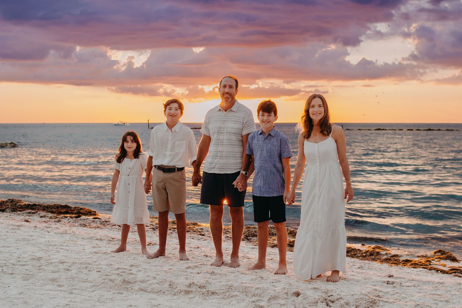 A group of people wearing white and blue outfits pose together on a sandy beach at sunset by the ocean.