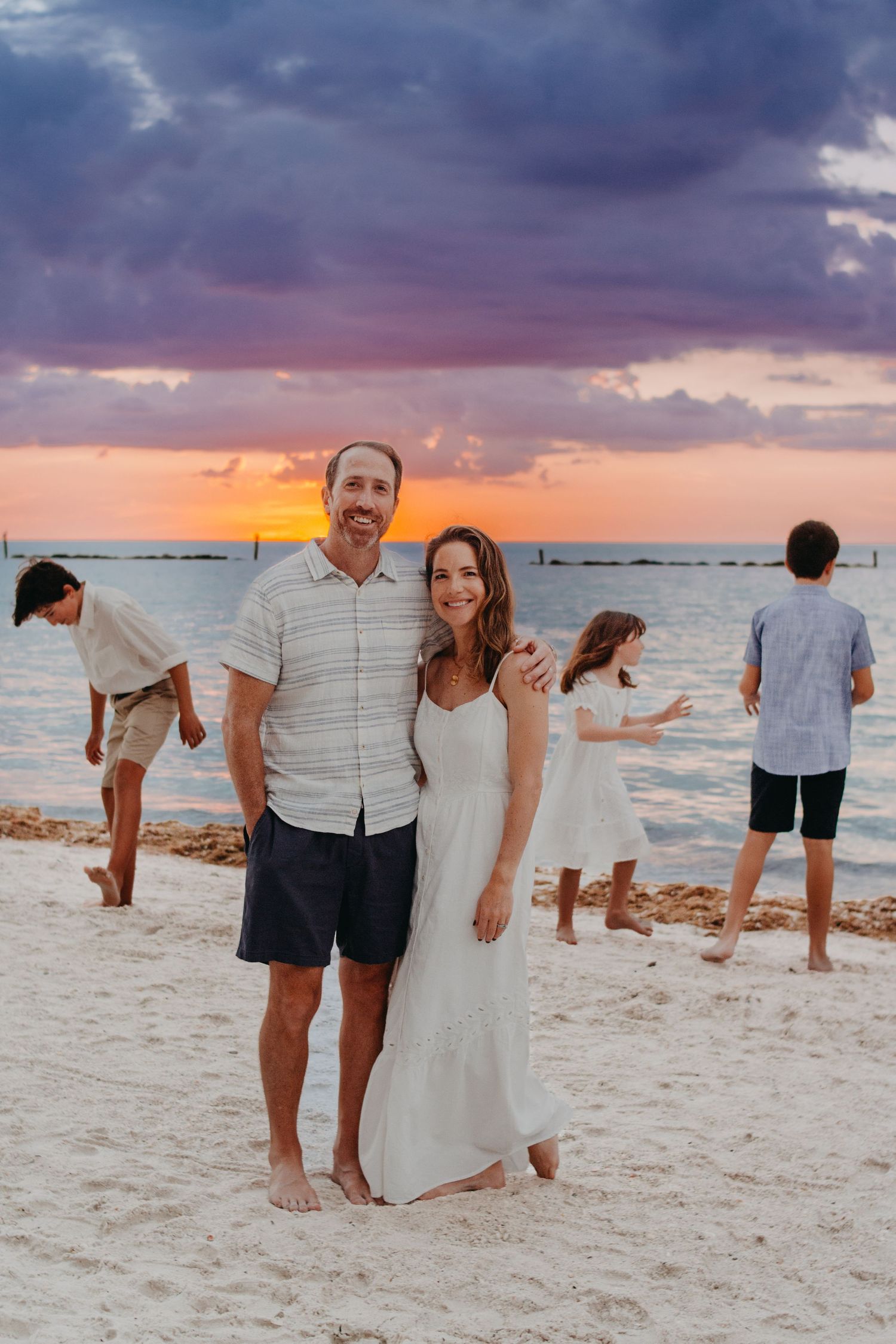 Beachgoers enjoy a stunning sunset on a sandy shore with dramatic purple and orange clouds over the ocean.