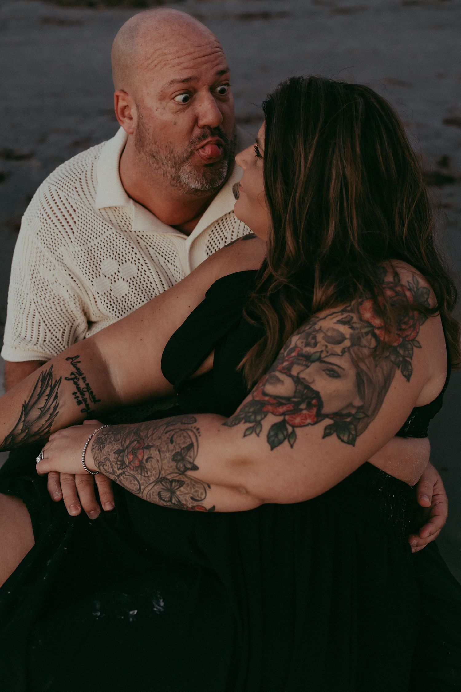 A romantic couple embraces on a dark beach, with one person wearing a white sweater and the other showing intricate tattoos.