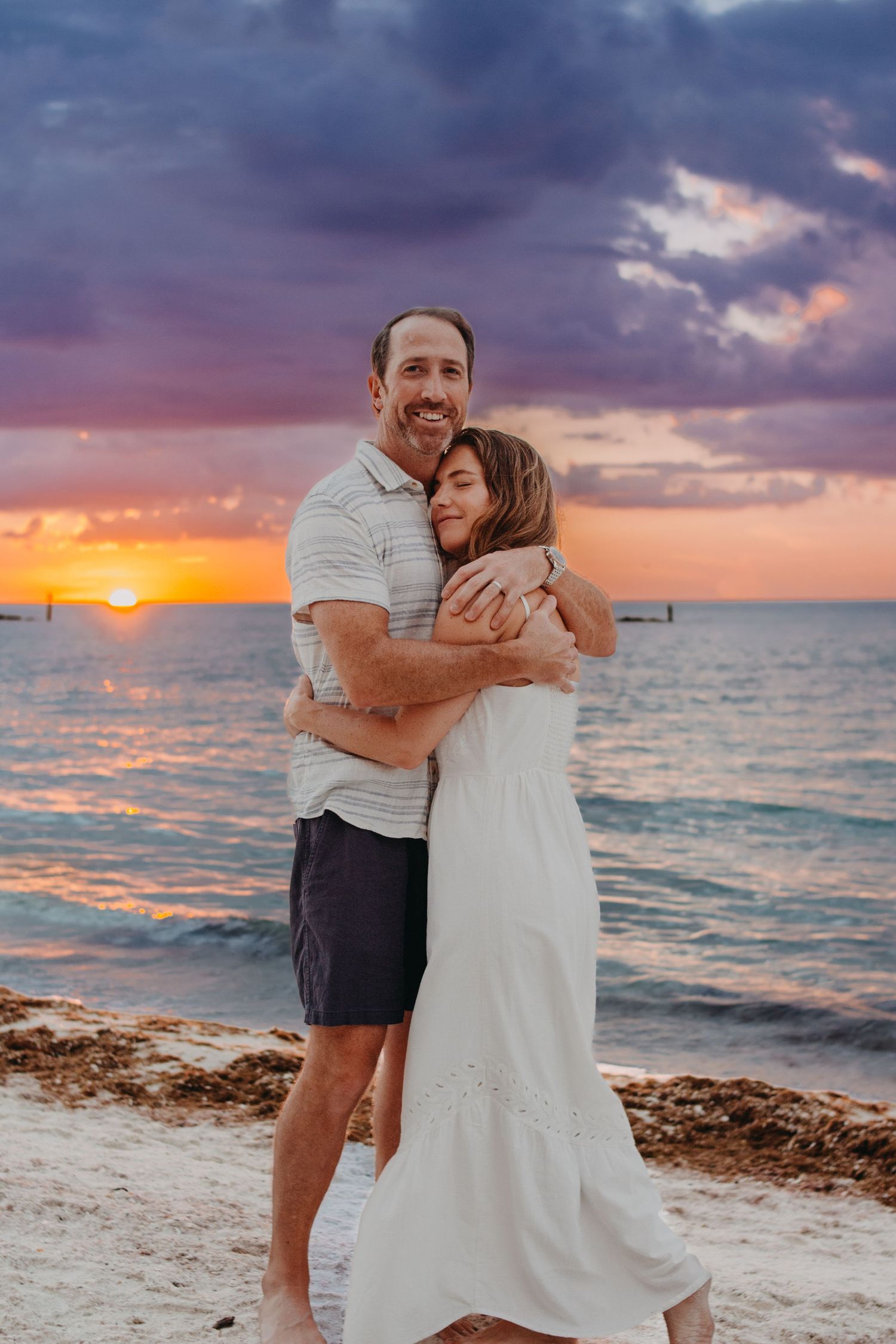 A couple embraces on a beach at sunset with dramatic purple and orange clouds reflecting on the ocean water.