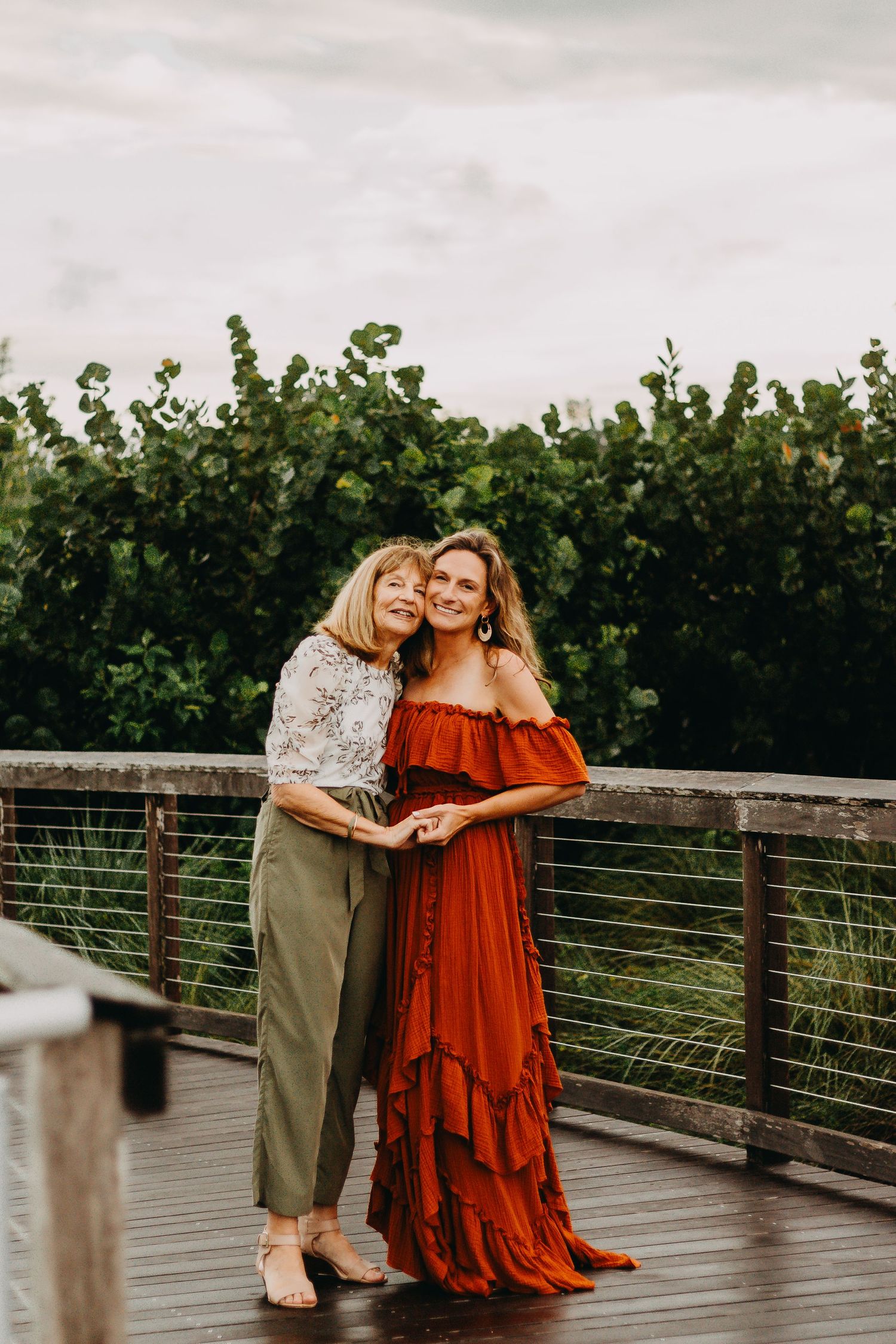 Two people embrace on a wooden deck surrounded by lush greenery, wearing a red flowy dress and casual outfit.
