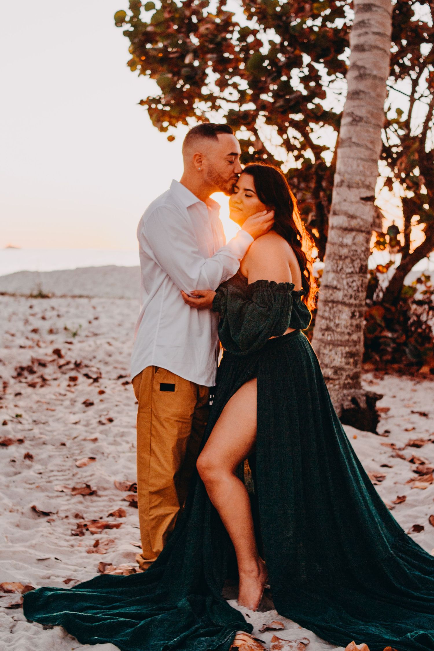 Romantic couple embraces on beach at sunset with woman wearing flowing emerald green dress and man in white shirt.