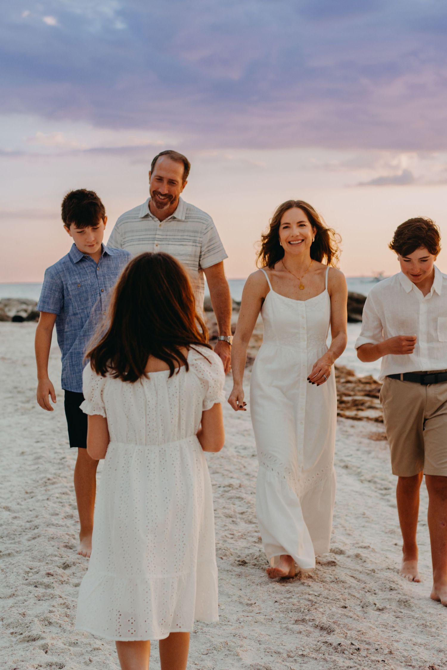 A family walks together on a sandy beach at sunset with soft pink and purple clouds in the sky.