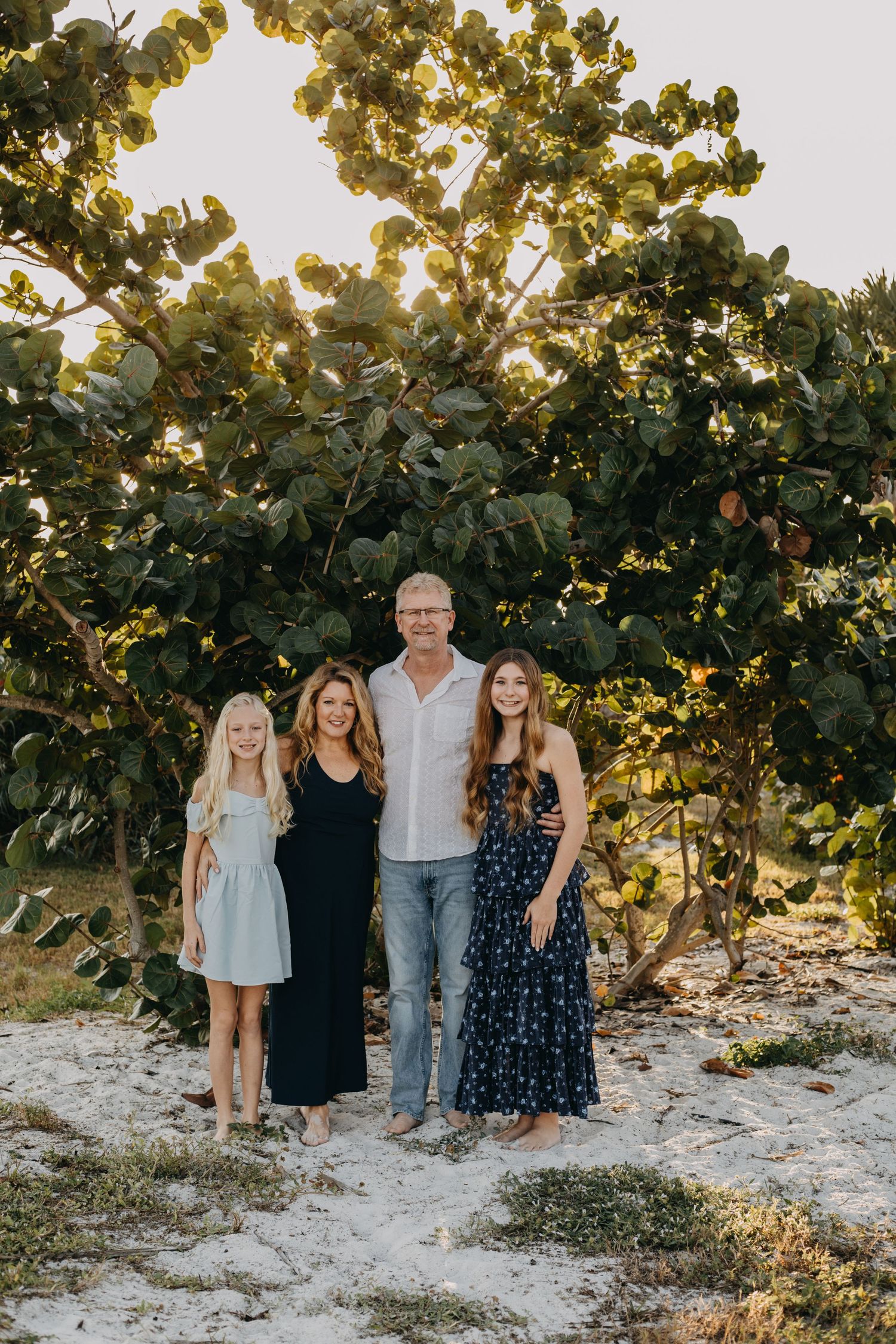 A group of four people stand together on a sandy beach path with lush green foliage and golden sunlight behind them.