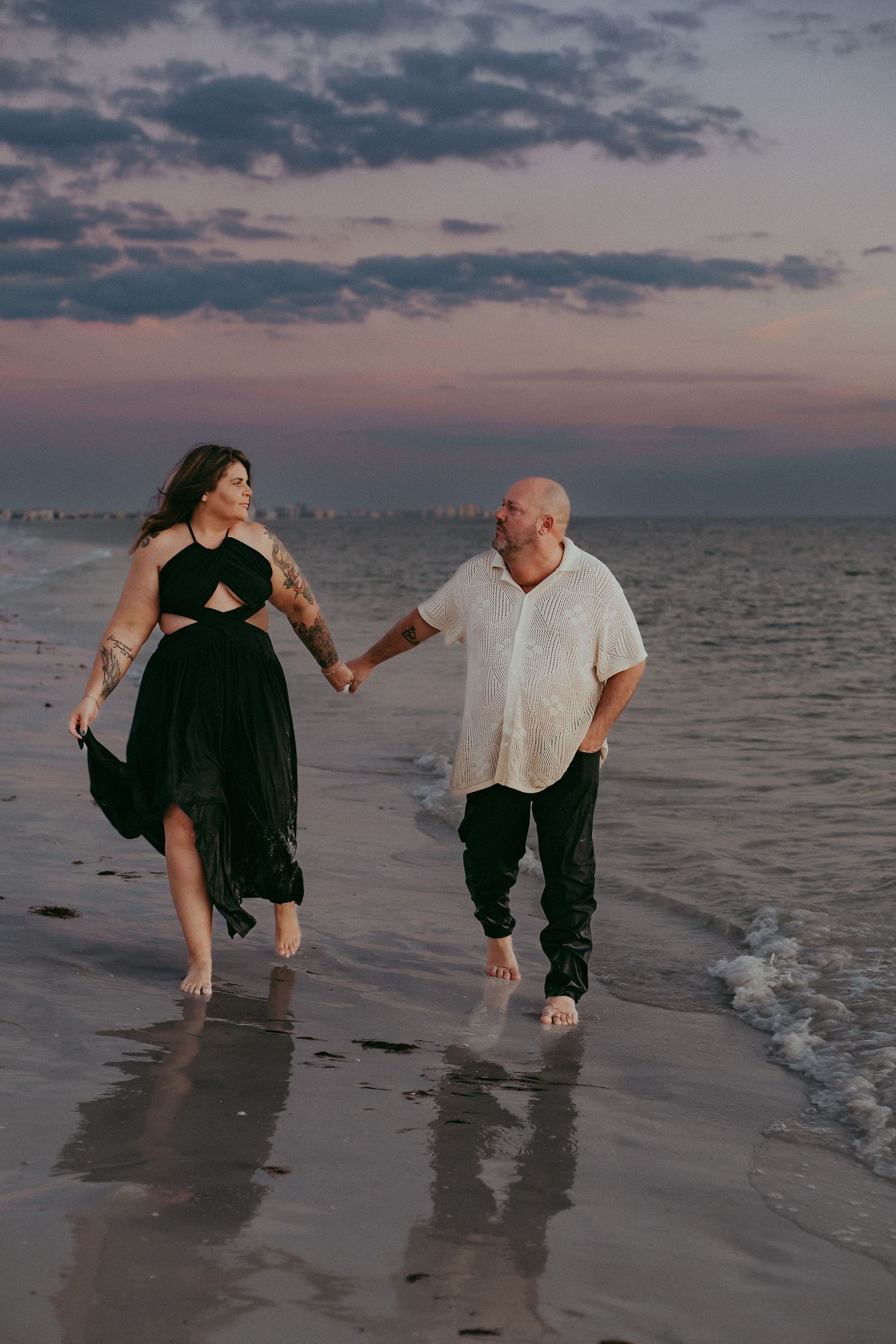 A couple walks hand in hand along a beach at sunset, with the woman in a black dress and man in white shirt.