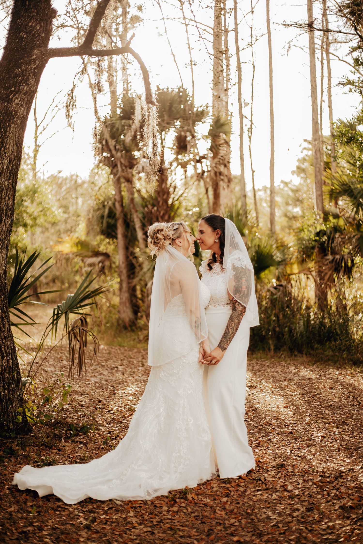 Two brides in white wedding dresses share an intimate moment on a sunlit forest path covered in fallen leaves.