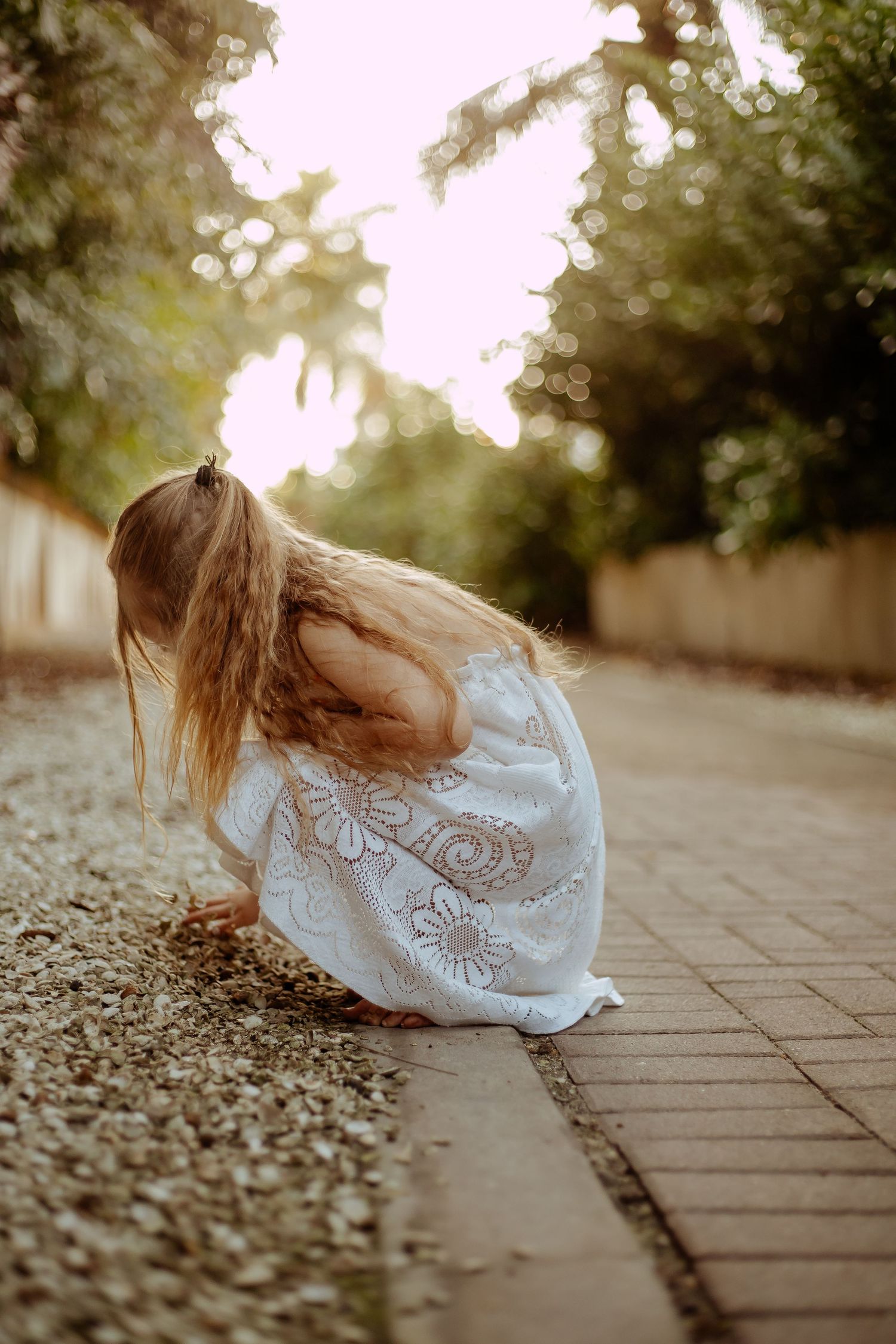 A person in a flowing white lace dress crouches down on a stone pathway during golden hour.