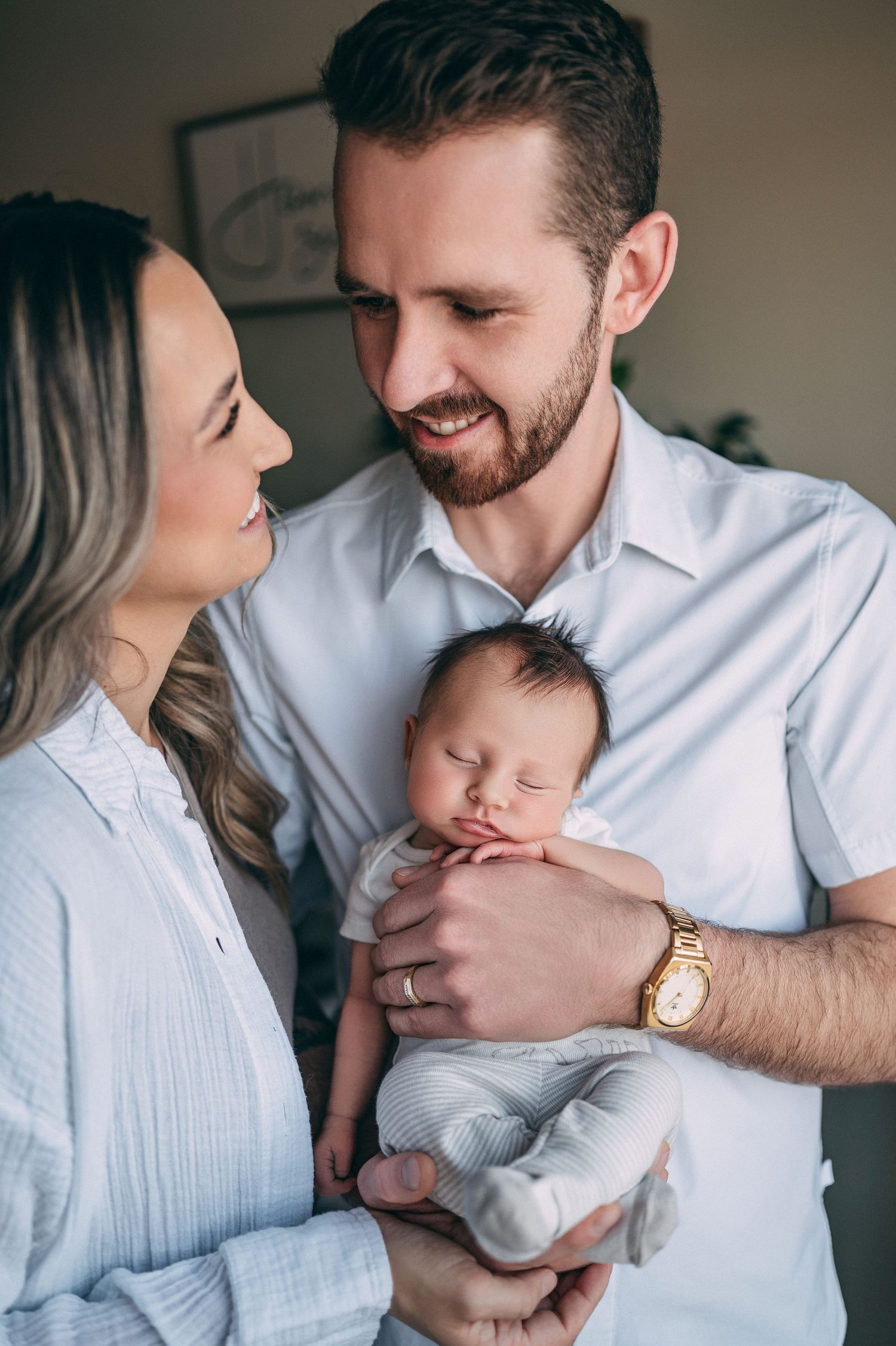 Parents sharing intimate moment while holding their sleeping newborn in white button-down shirts.