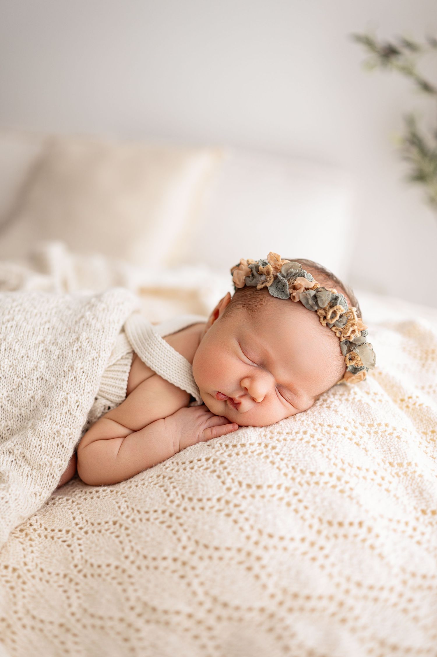 A sleeping newborn baby wearing a floral headband rests peacefully on a white textured blanket in soft natural light.