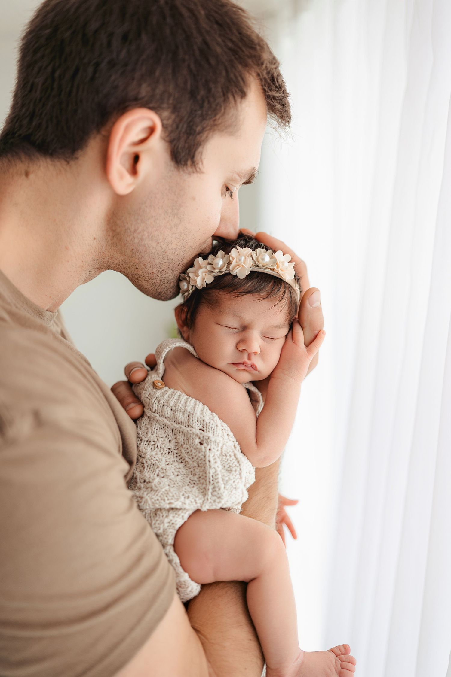 A parent wearing tan clothing holds a sleeping baby in a lace dress against a white curtain background.