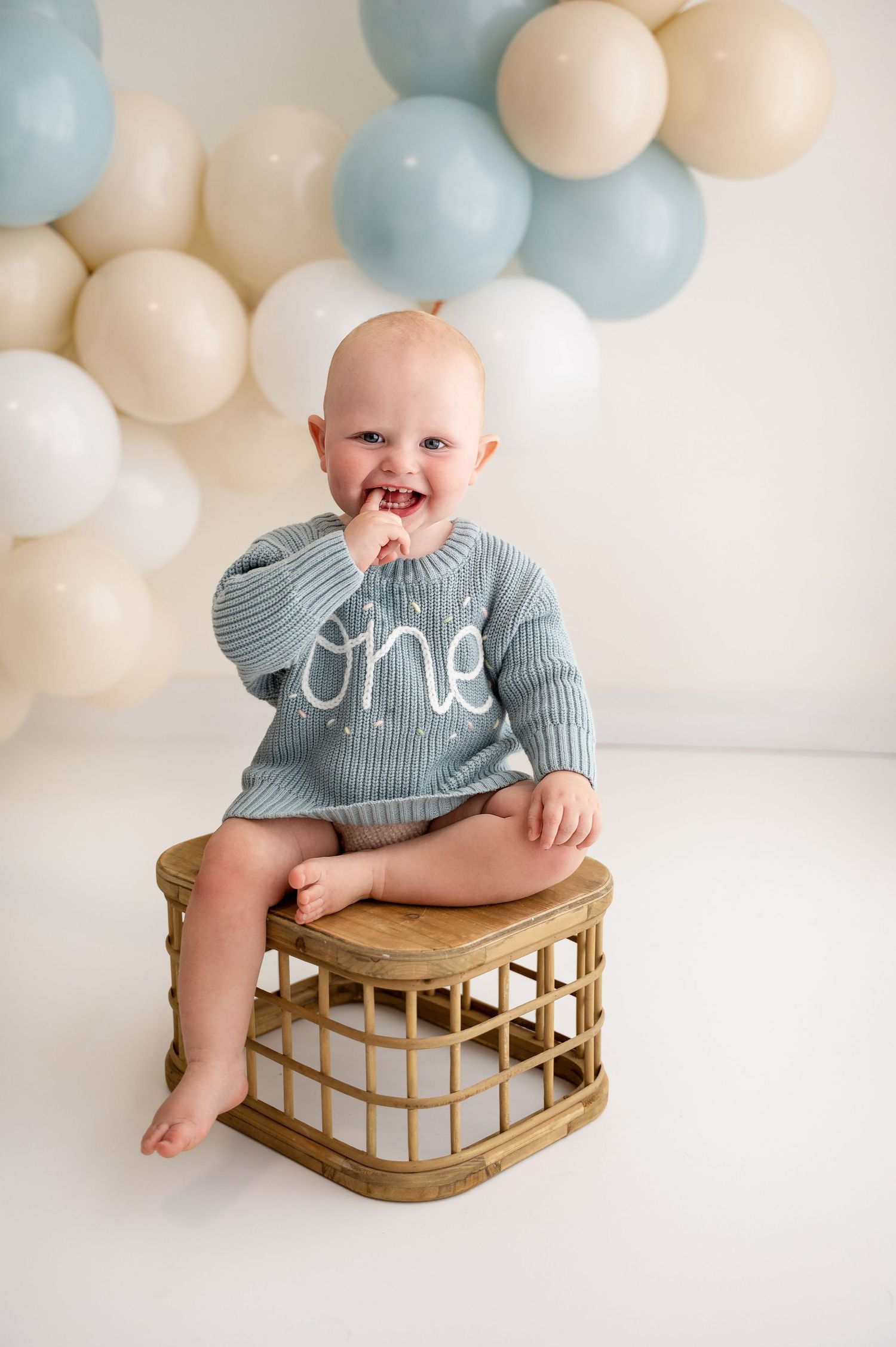 Baby in gray sweater sits on wooden stool with balloon decorations in background.
