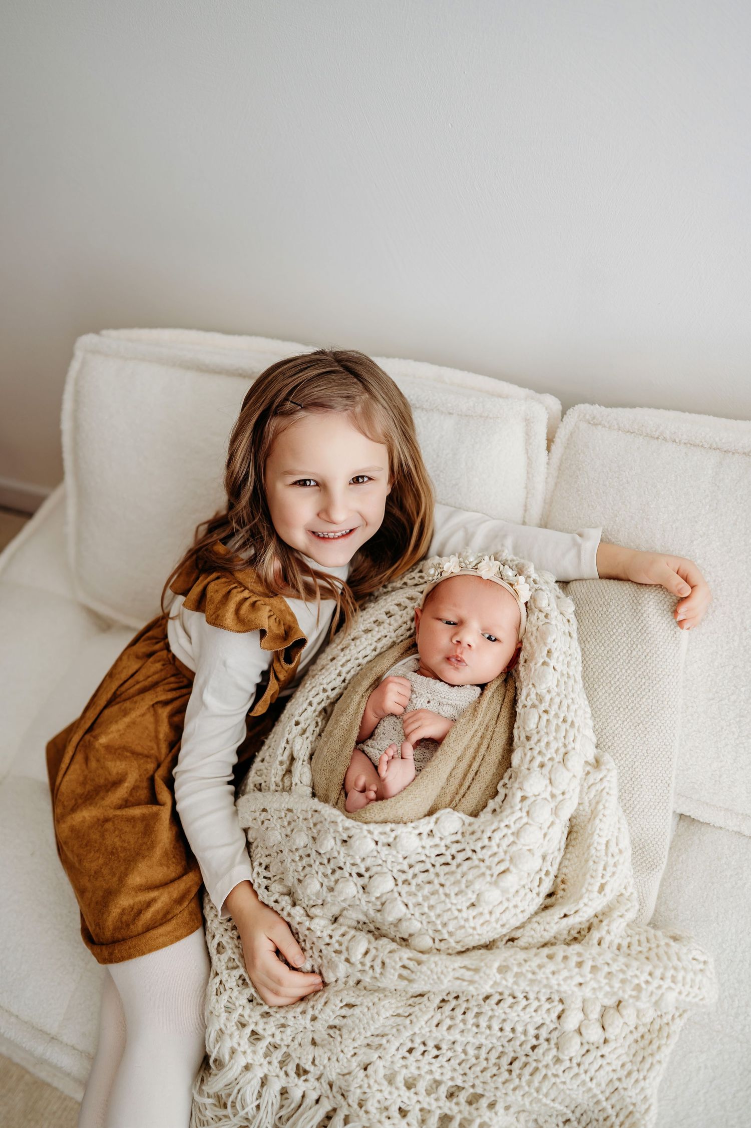 Two children sit together on a white couch with one wrapped in a chunky knit cream-colored blanket.