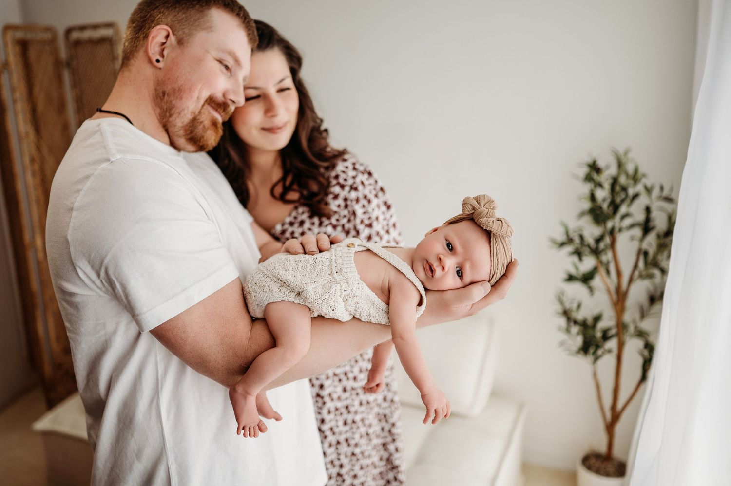 A tender family moment indoors as parents cuddle their sleeping baby in a serene white room with greenery.