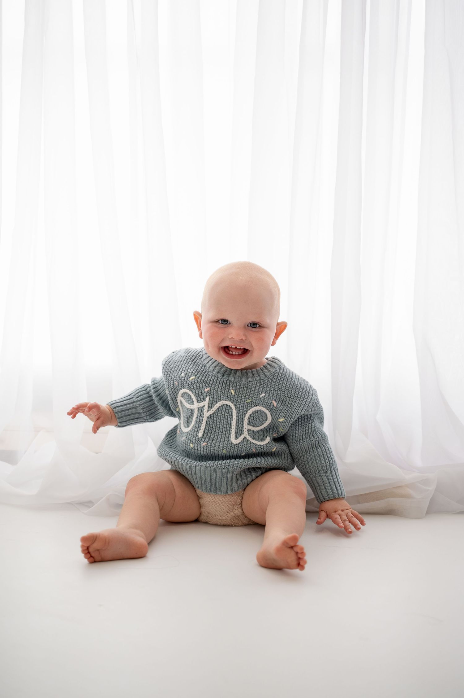 Baby in gray sweater crawls and smiles on white floor in bright studio setting.