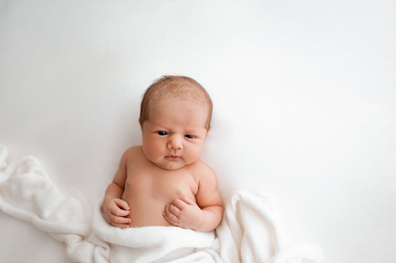 A newborn baby lies peacefully on soft white bedding during a professional photography session.