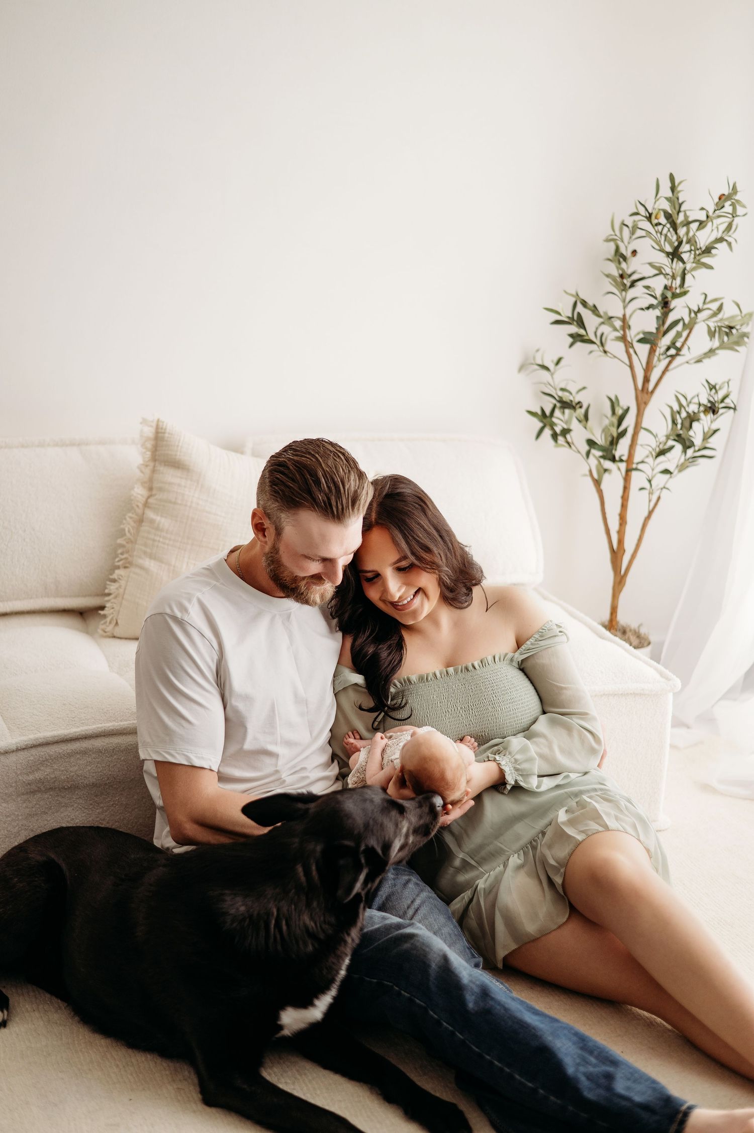 A couple relaxes on the floor with their black dog in a bright, minimalist room with a small potted tree.