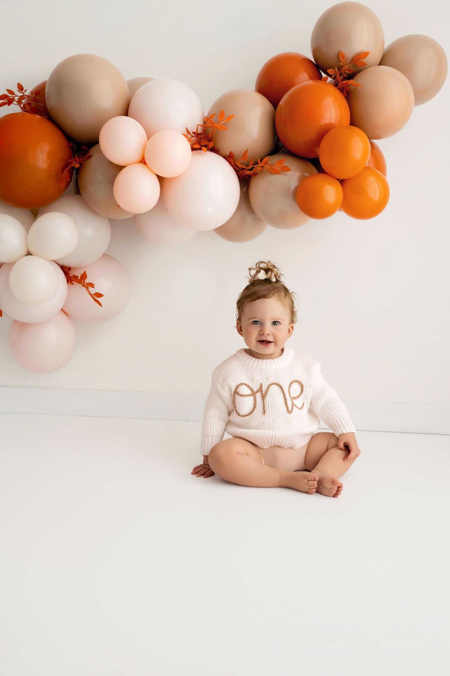 Baby in white onesie sits on floor with peach and orange balloon arch backdrop for first birthday photoshoot.