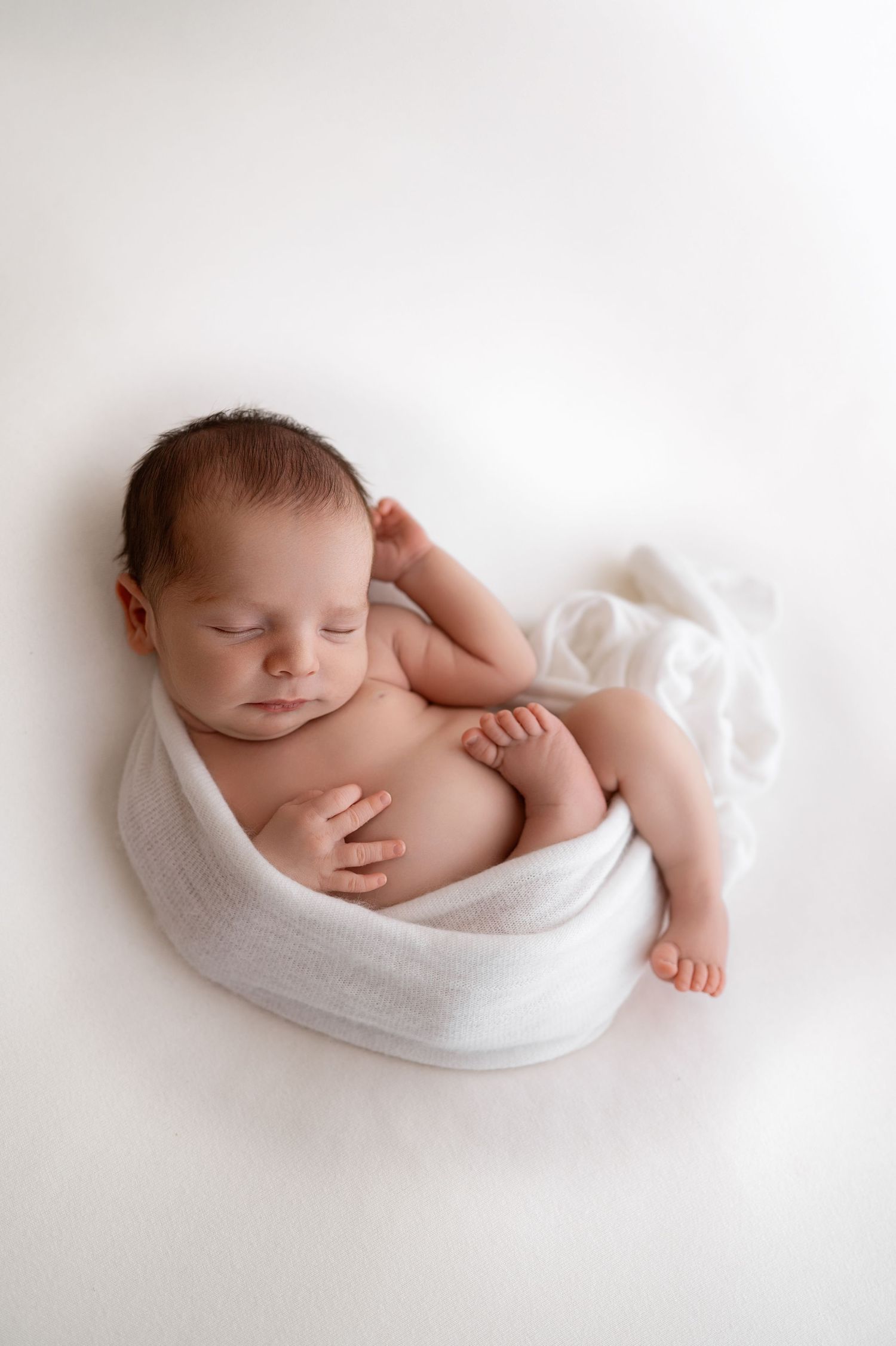 Sleeping newborn swaddled in white fabric on clean white backdrop.