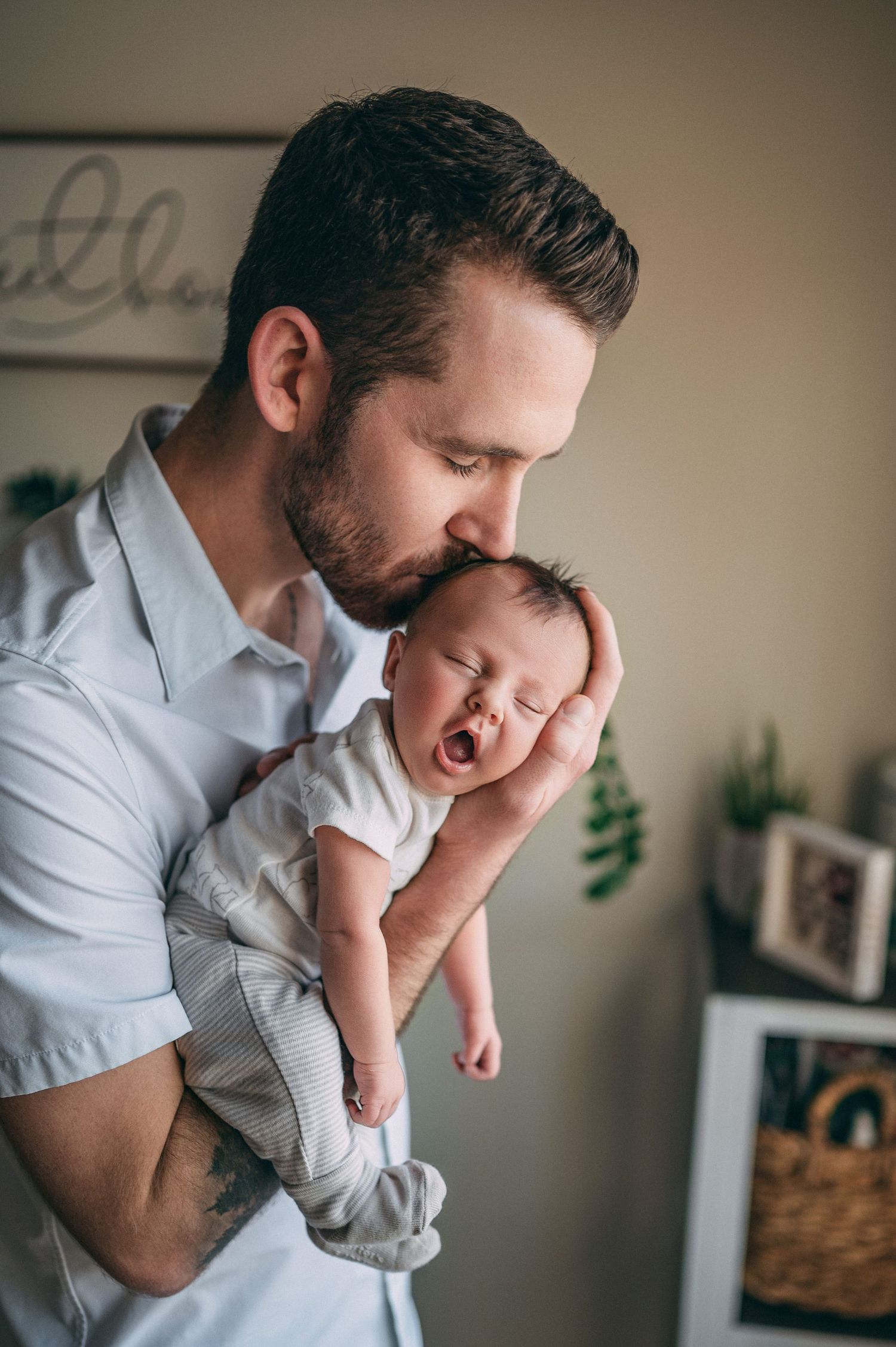 Parent in white shirt holding and kissing sleeping newborn baby against brown wall with decor.