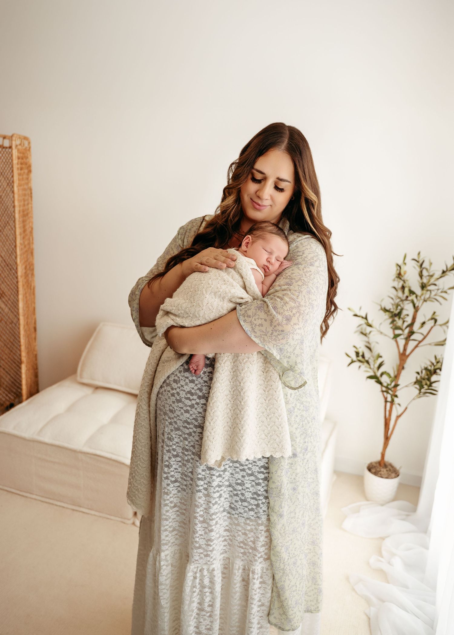 Woman in white lace dress holding a baby while standing in a bright minimal bedroom interior with potted plant.