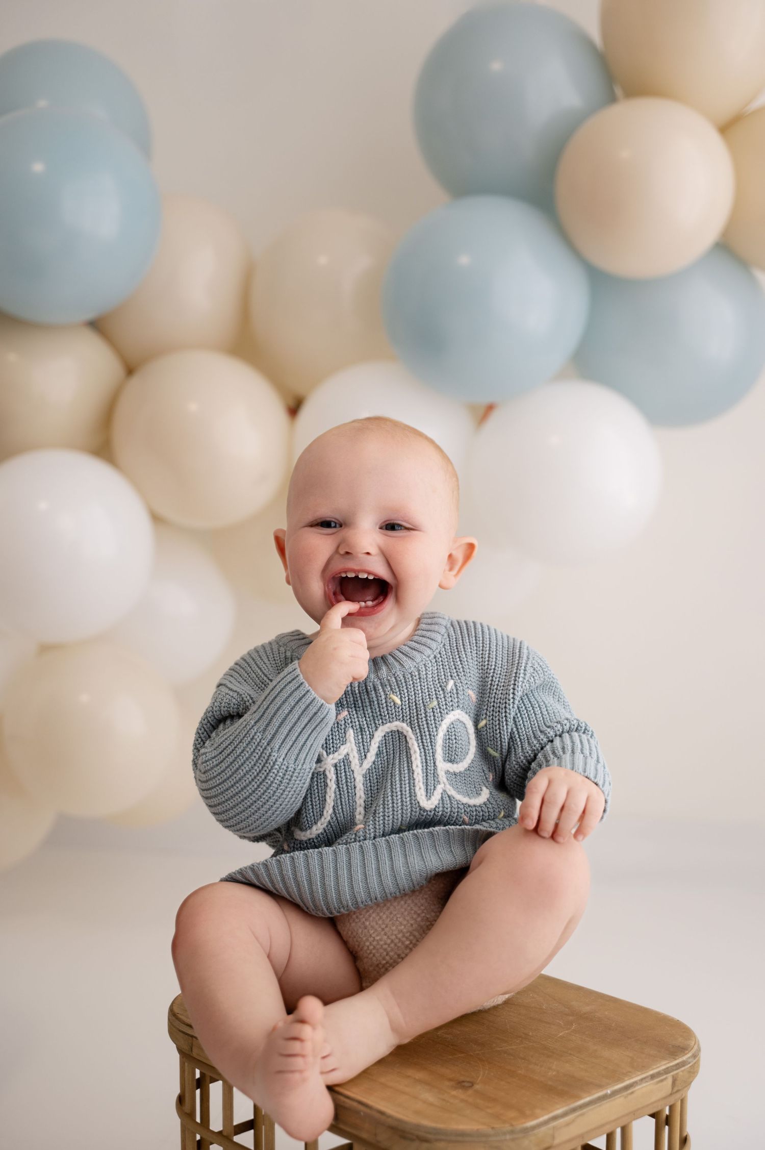 Baby wearing gray striped onesie sits on wooden stool against white and blue balloon backdrop.