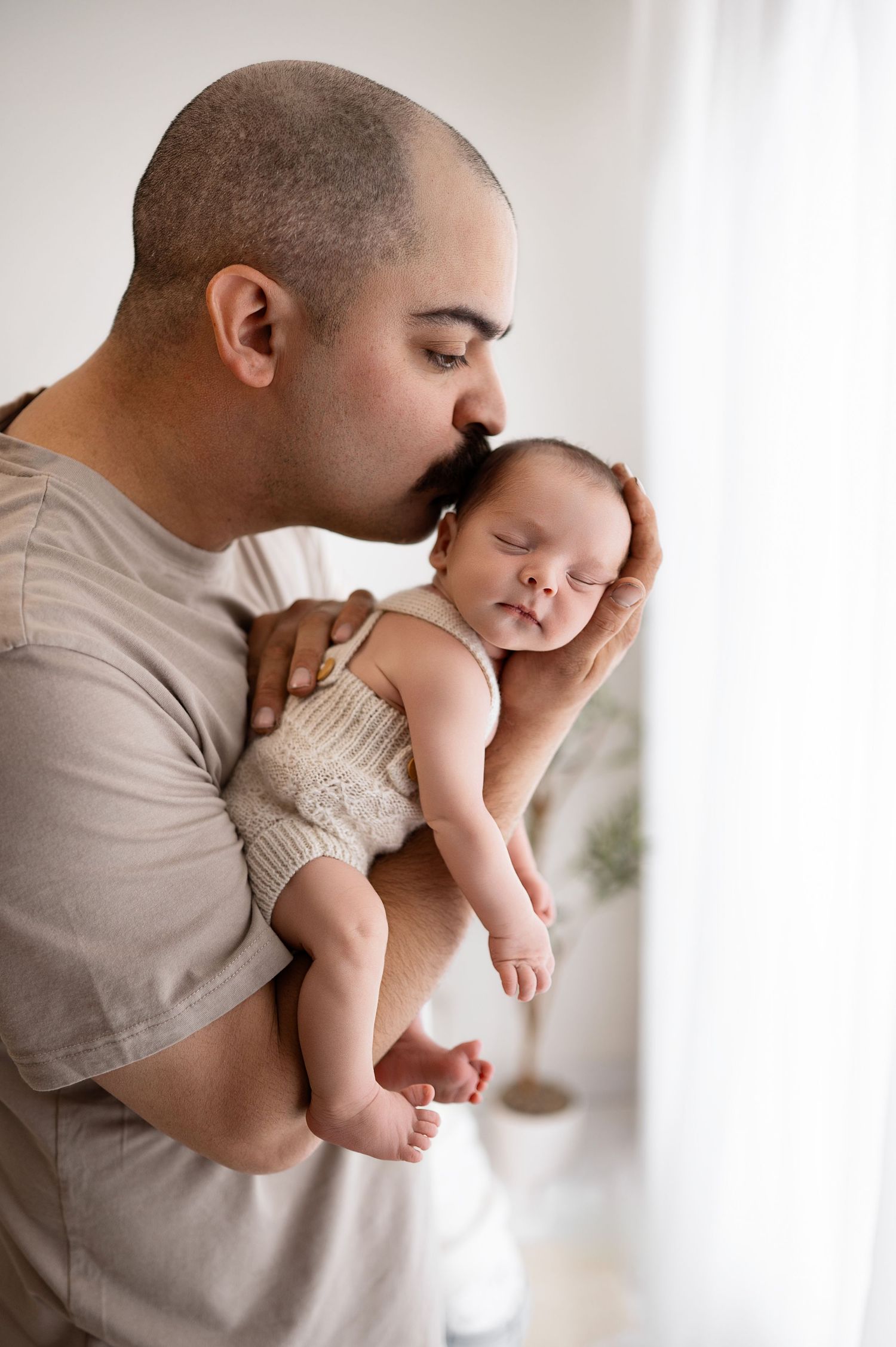A parent tenderly holds and kisses a baby wearing a knit romper in a bright, minimalist setting.