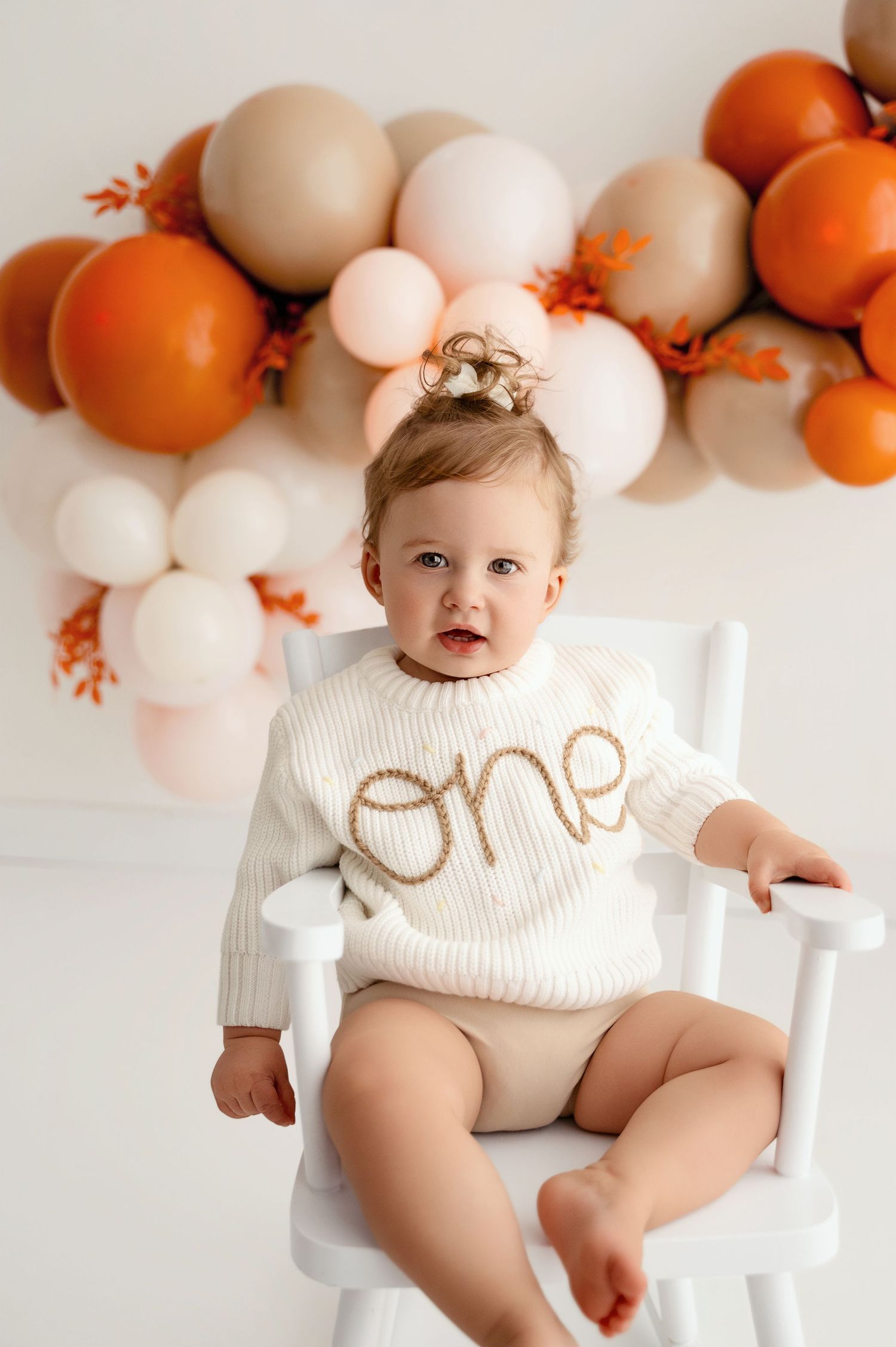 White and orange balloon garland decorates a minimalist first birthday photo session against a white backdrop.