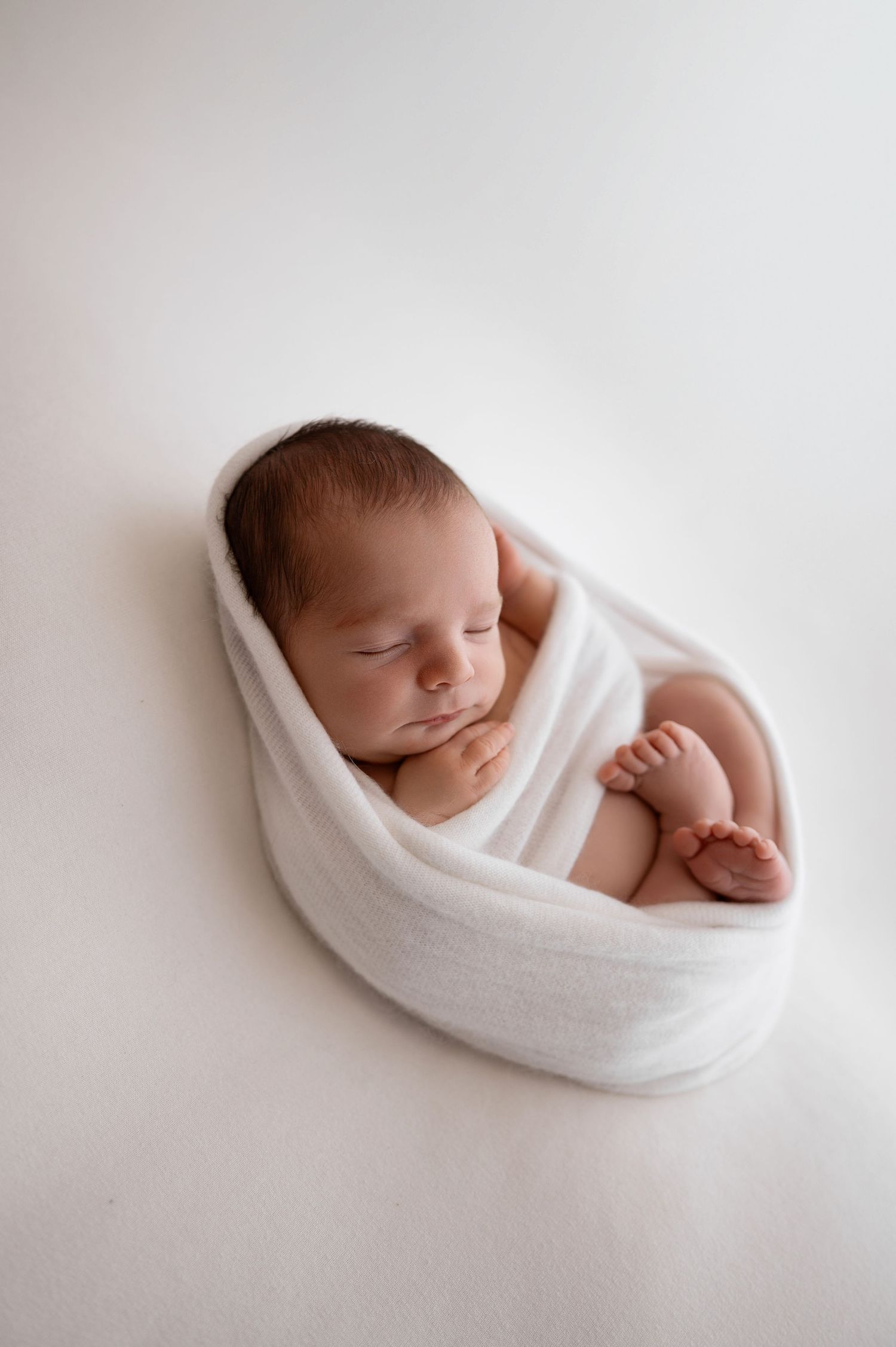 Peaceful newborn baby wrapped in soft white blanket against minimalist white background.