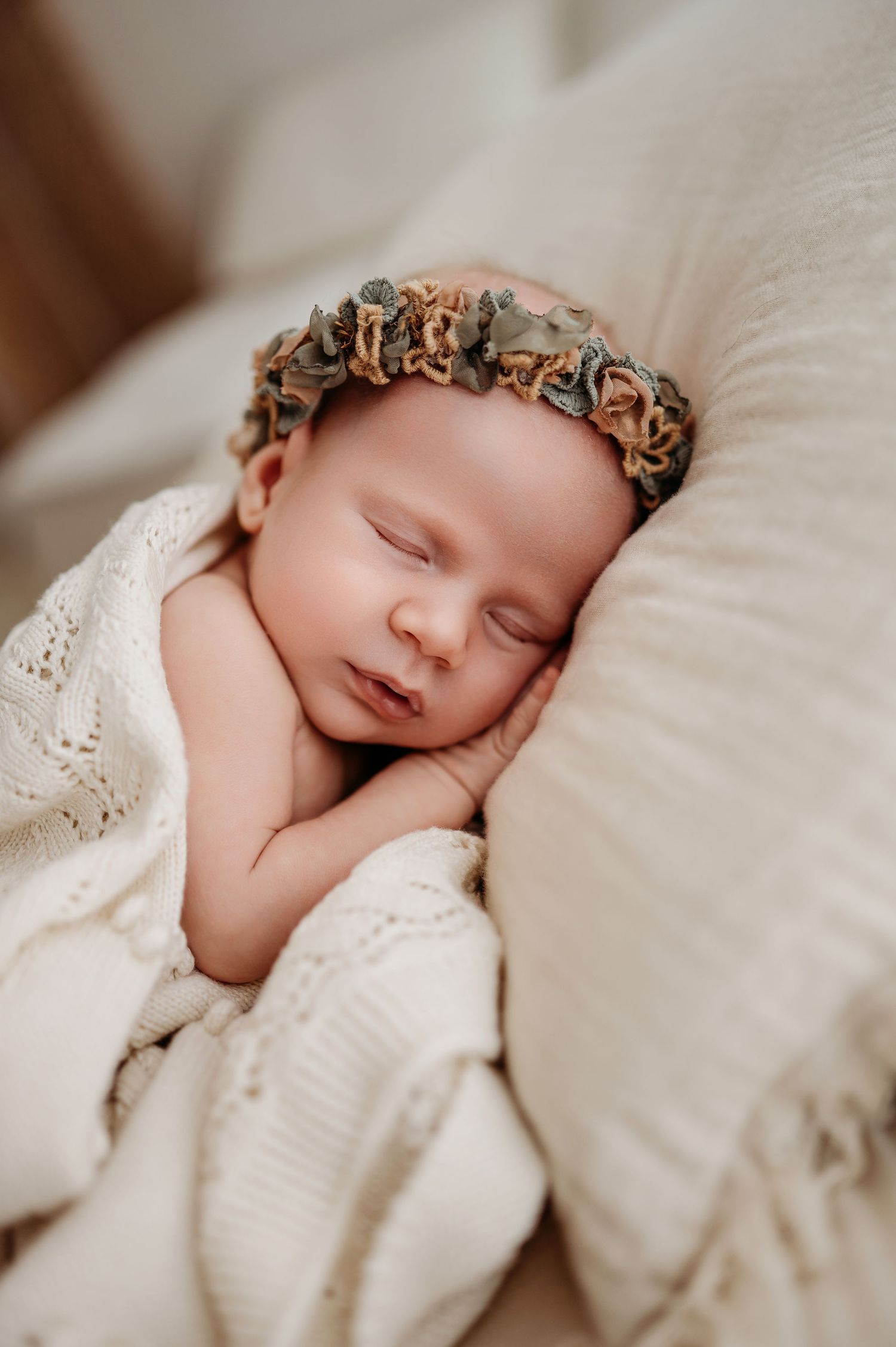 Sleeping newborn wearing floral headband wrapped in cozy white knit blanket during professional photo session.