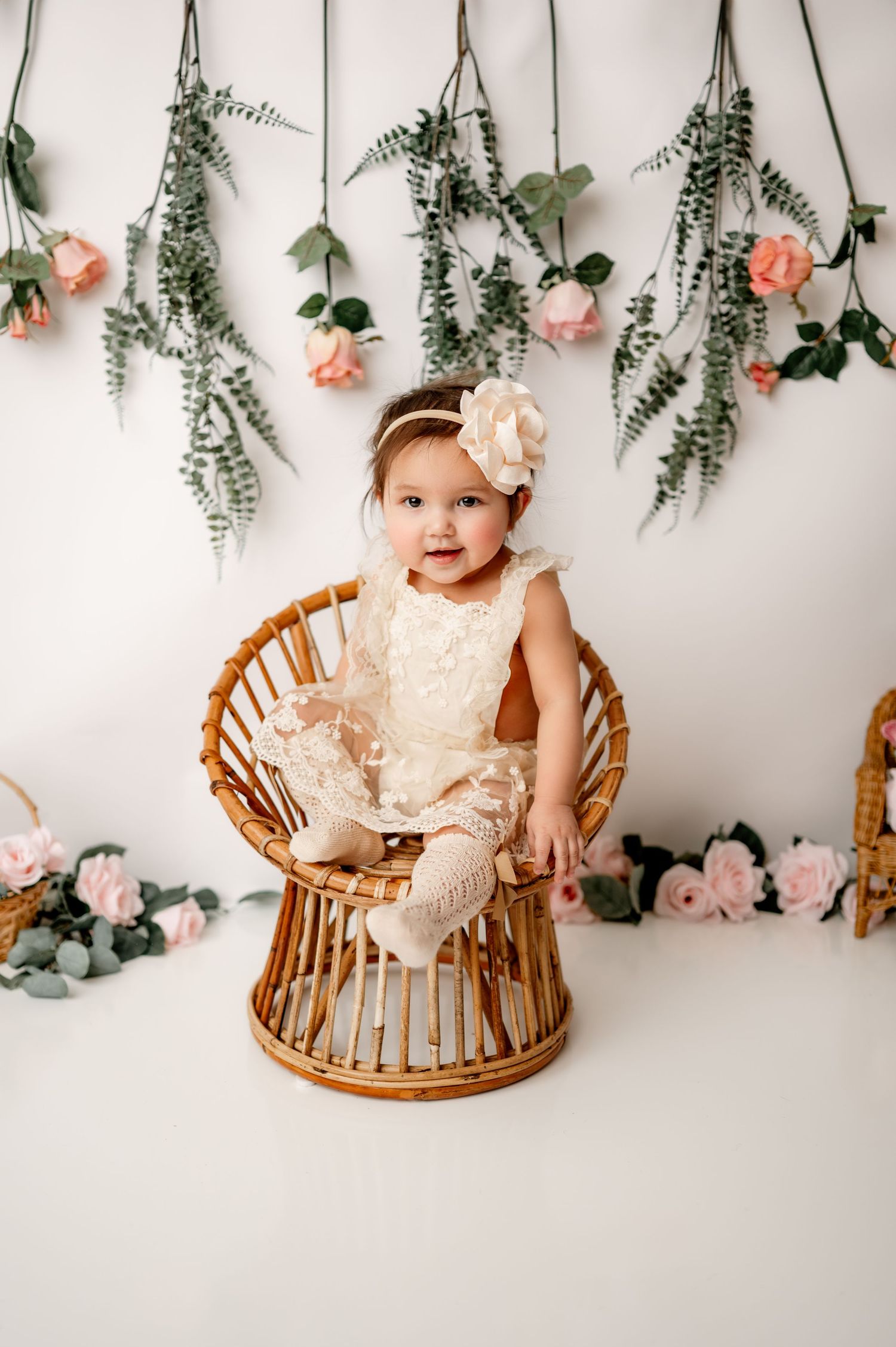 Rattan chair sits beneath a romantic floral backdrop with hanging eucalyptus and peach roses against white wall.