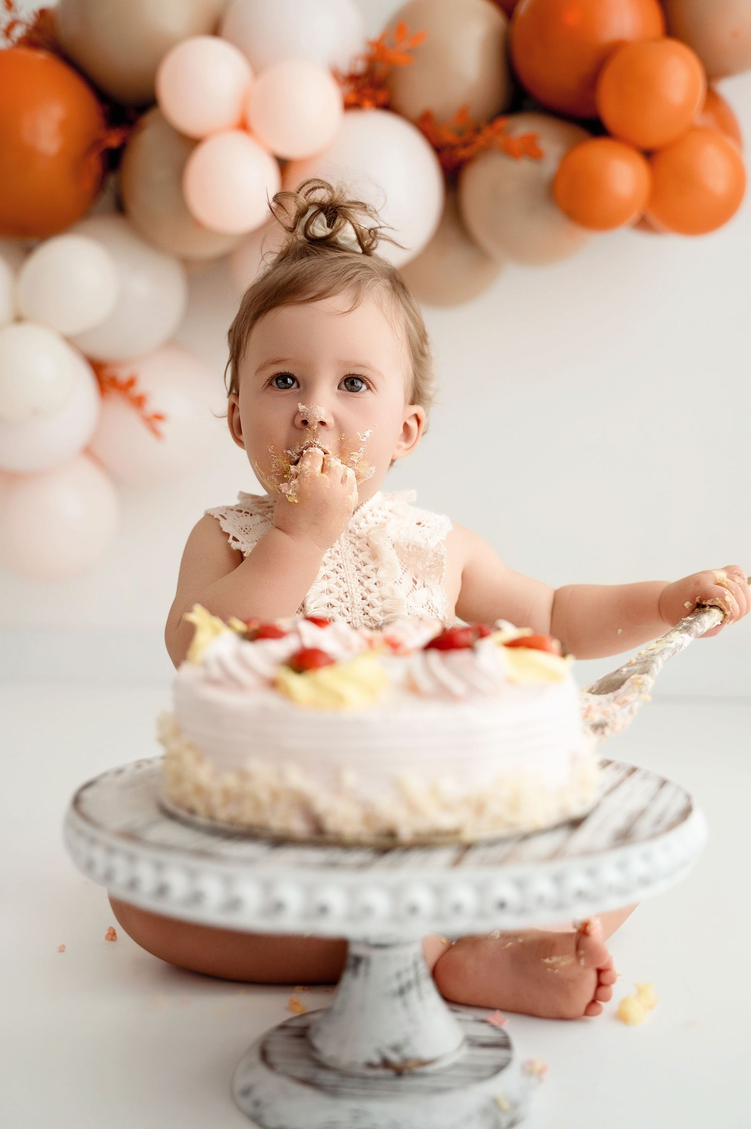 Baby enjoys messy cake smash photography session with white and peach balloon backdrop on vintage cake stand.