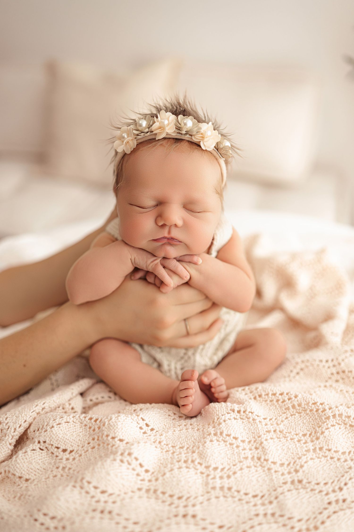 A sleeping newborn baby wearing a floral crown lies peacefully on soft lace bedding in a warm natural light setting.