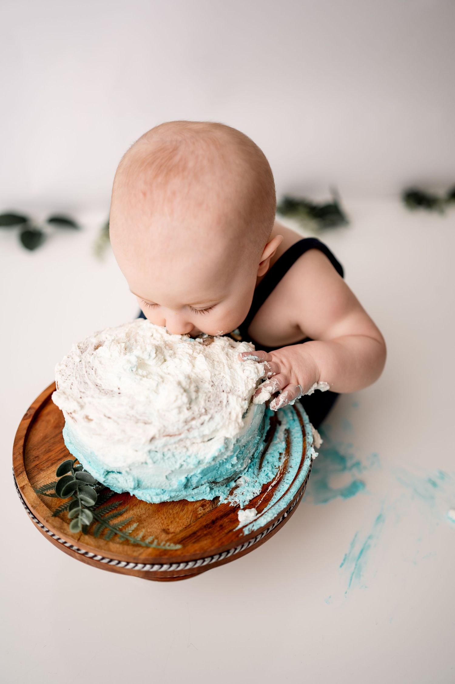 Baby in a cake smash photo session dives face first into a blue and white frosted cake on a wooden plate.