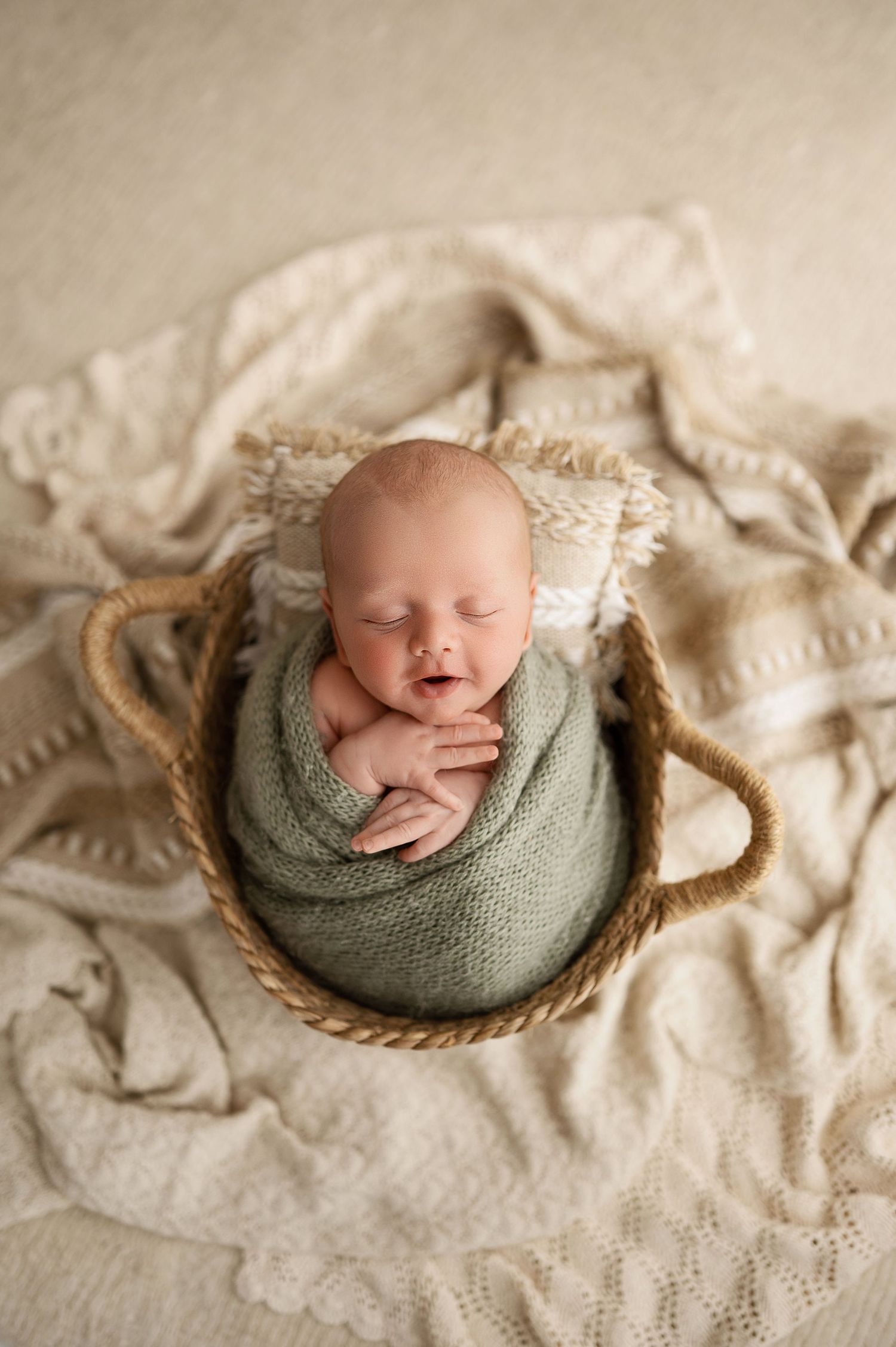 A sleeping newborn swaddled in sage green wrap lies peacefully in a woven basket on cream colored textured fabric.