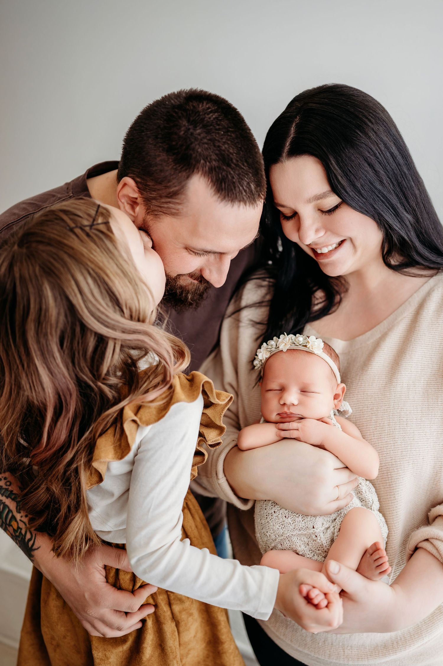 A loving family moment with parents and young child gazing adoringly at their sleeping newborn baby wearing a floral headband.