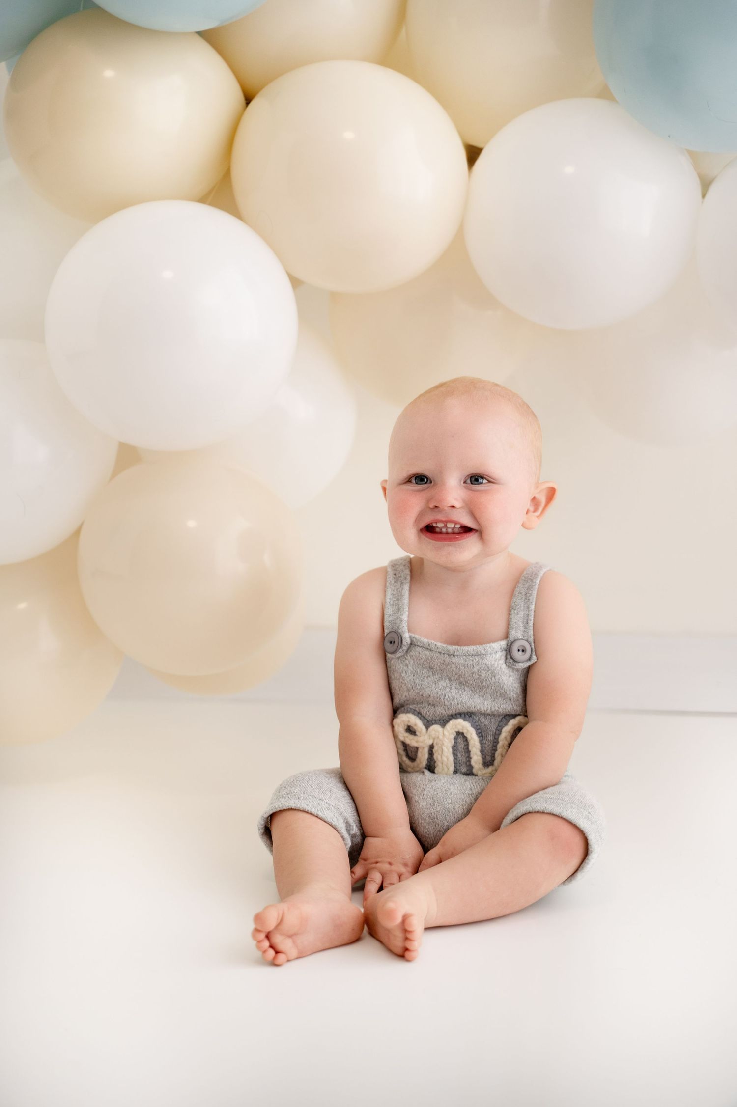 A baby wearing gray overalls sits on a white floor against a backdrop of cream-colored balloons.