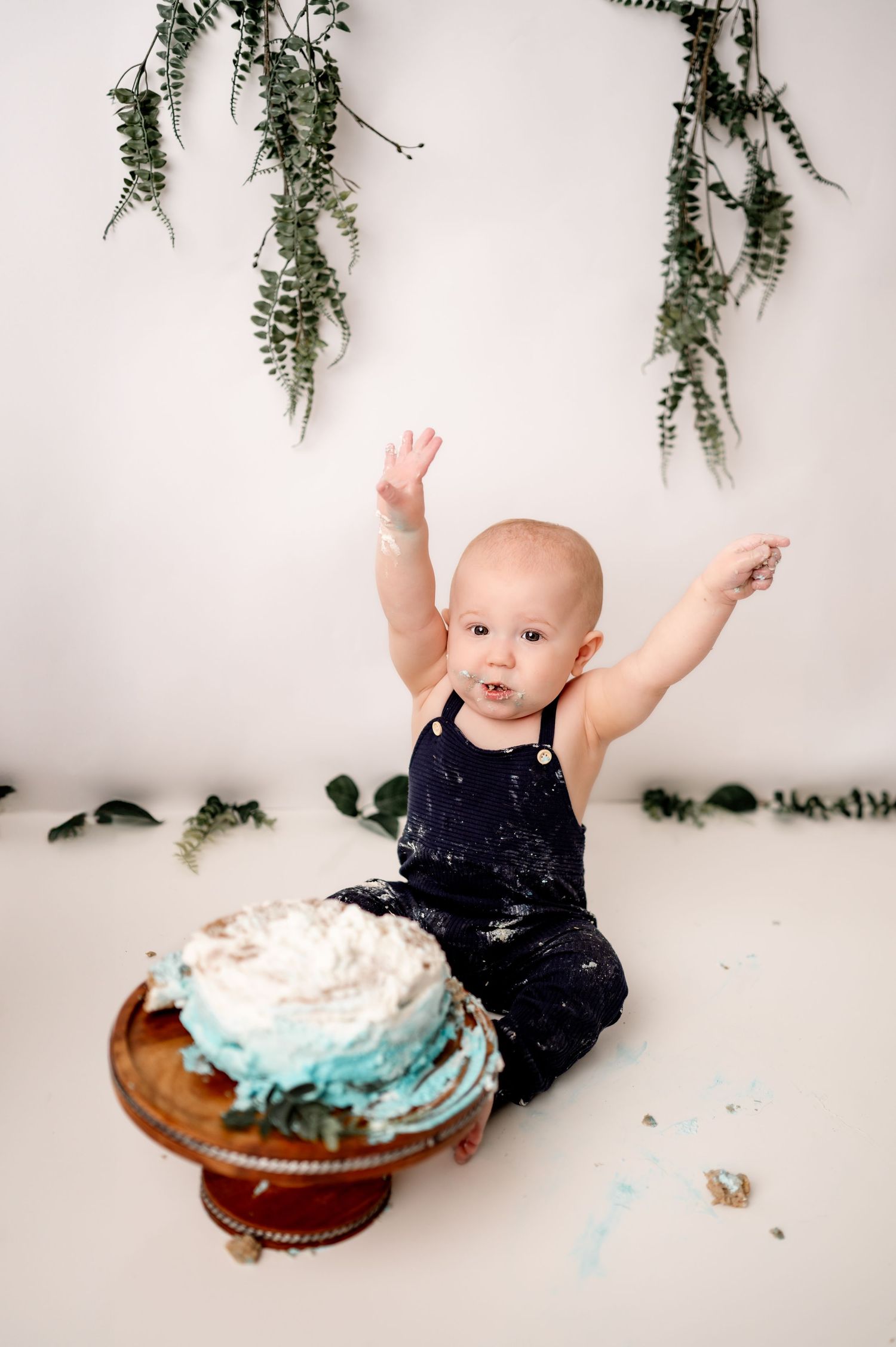 Baby wearing navy outfit reaches excitedly for a smash cake during a first birthday photoshoot with greenery.